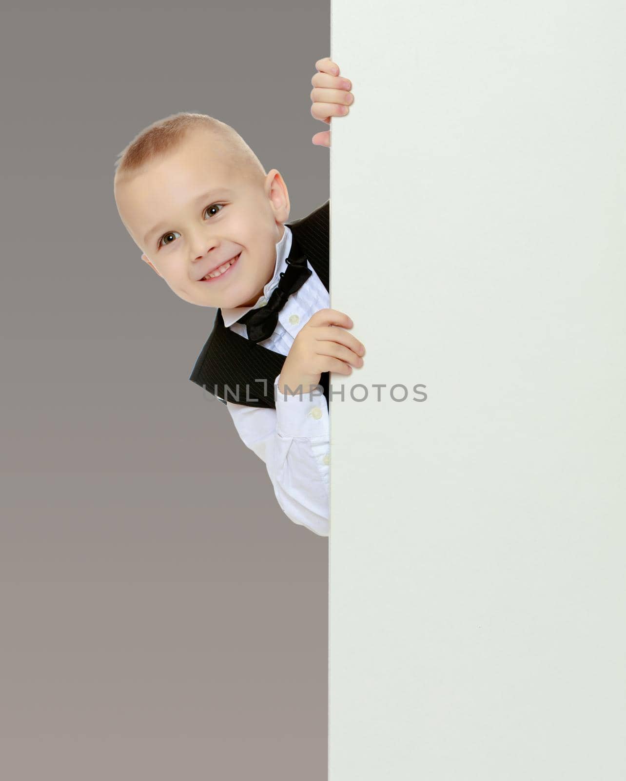 Cute little blonde boy in a black waistcoat and a white shirt and a bow at the neck , peeping over white banner.On a gray background.