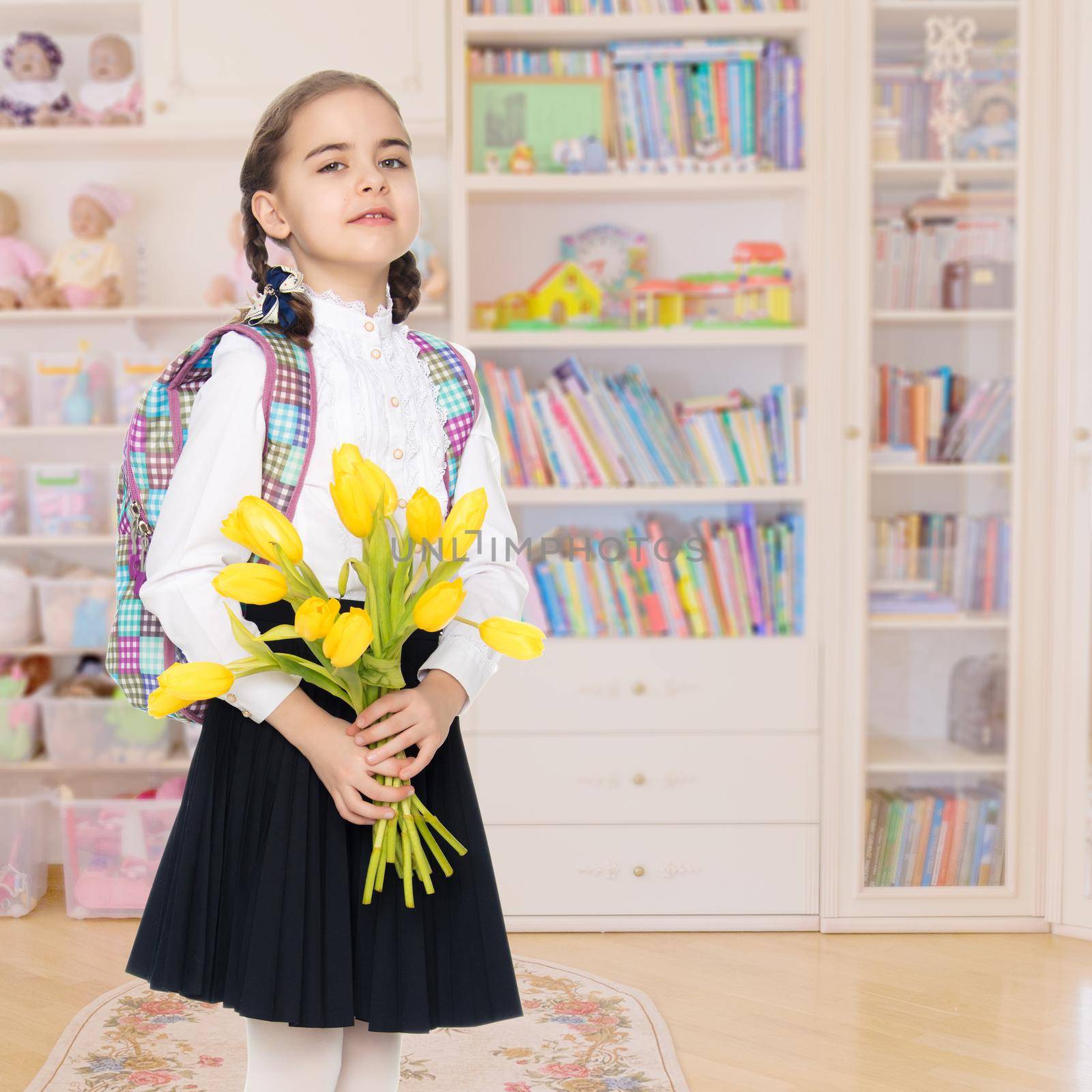 Beautiful little blond schoolgirl, with long neatly braided pigtails. In a white blouse and a long dark skirt.She is holding a bouquet of yellow tulips.In the children's room.