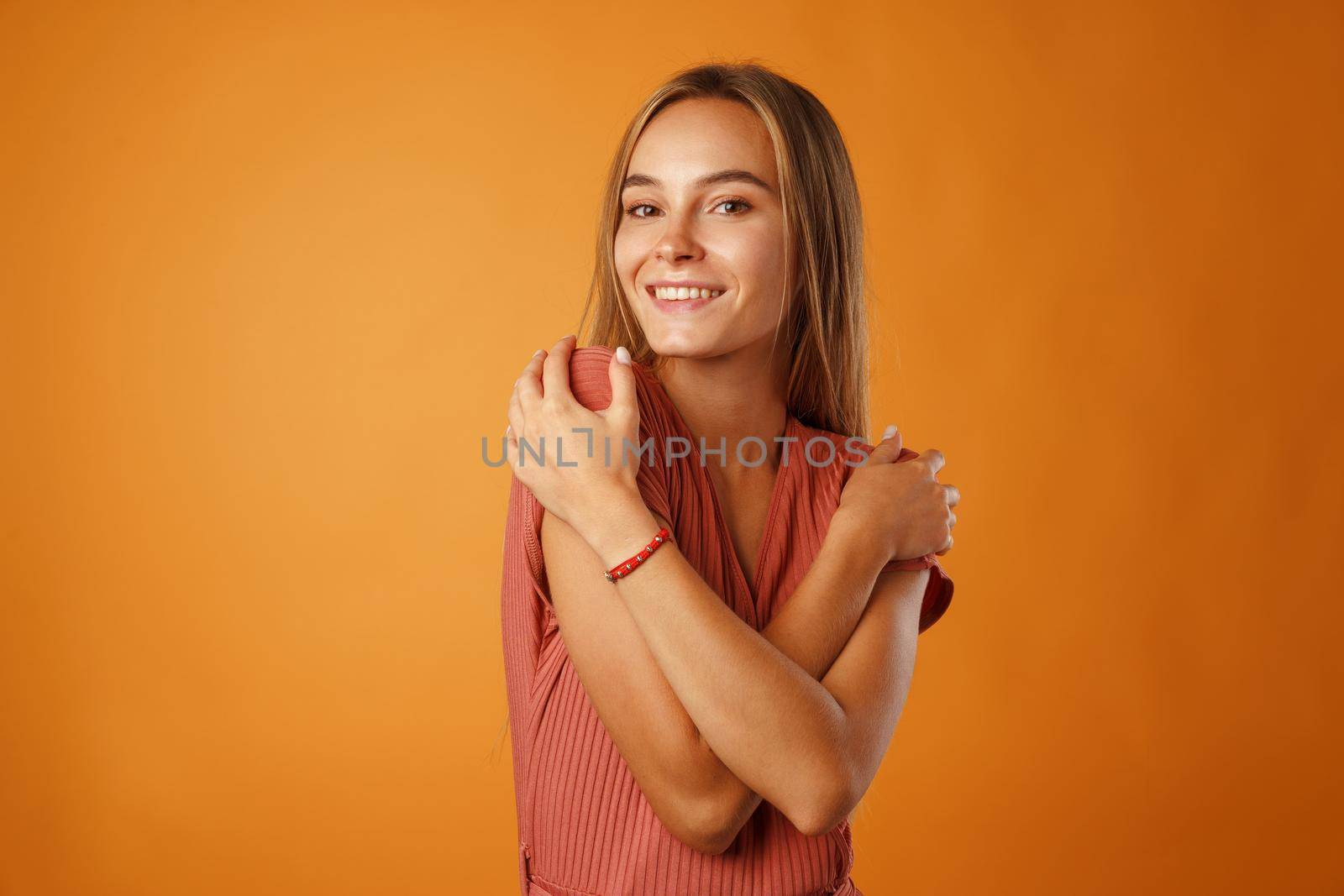 Peaceful young blonde woman holding hands on her chest against orange background.