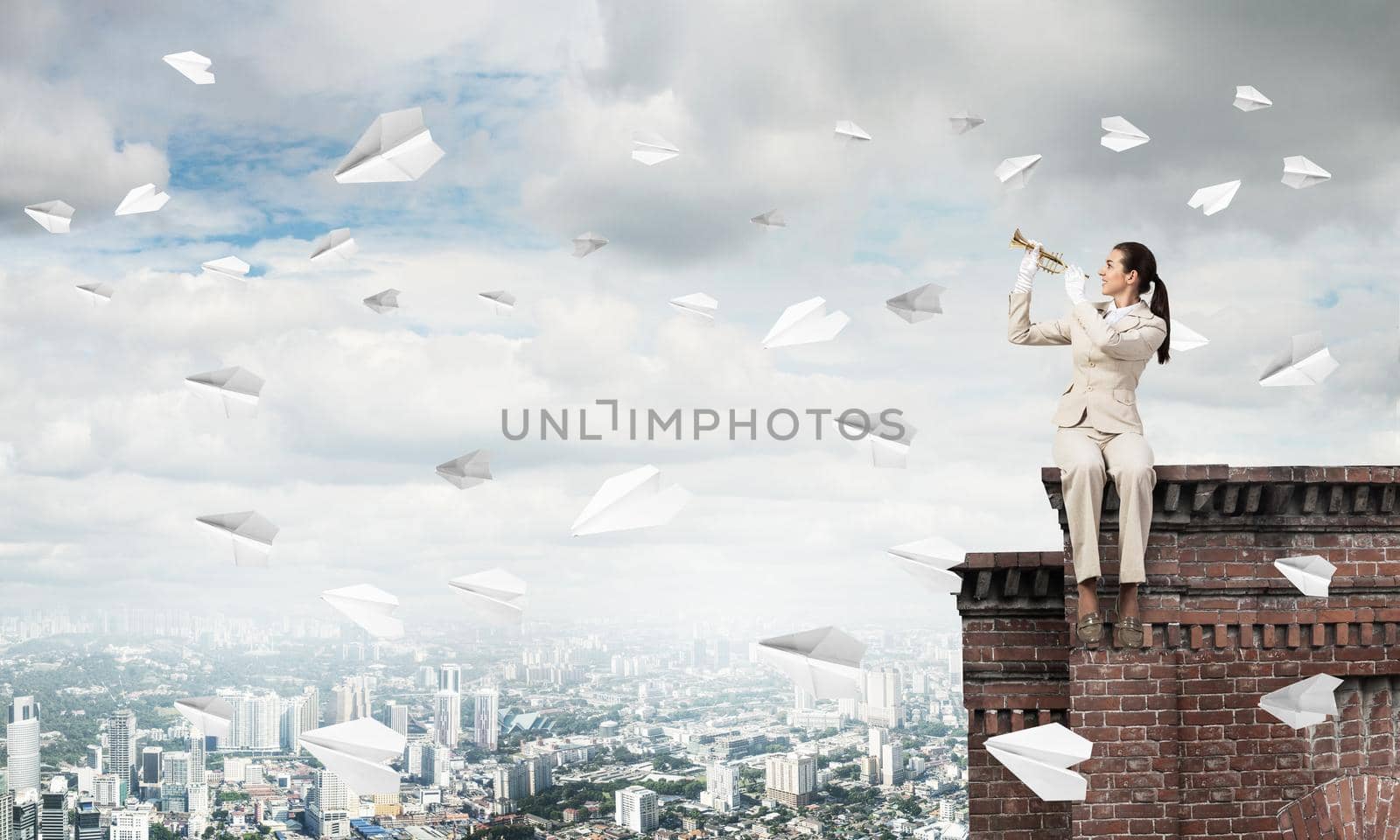Young woman playing trumpet and sitting on roof. Girl in white business suit with music brass instrument posing high above modern city. Imagination and inspiration. Flying paper planes in blue sky