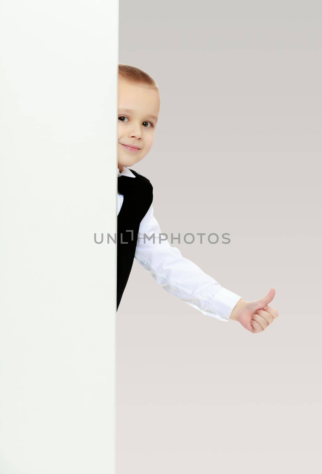 Cute little blonde boy in a black waistcoat and a white shirt and a bow at the neck , peeping over white banner.He holds up a thumb.On a gray background.