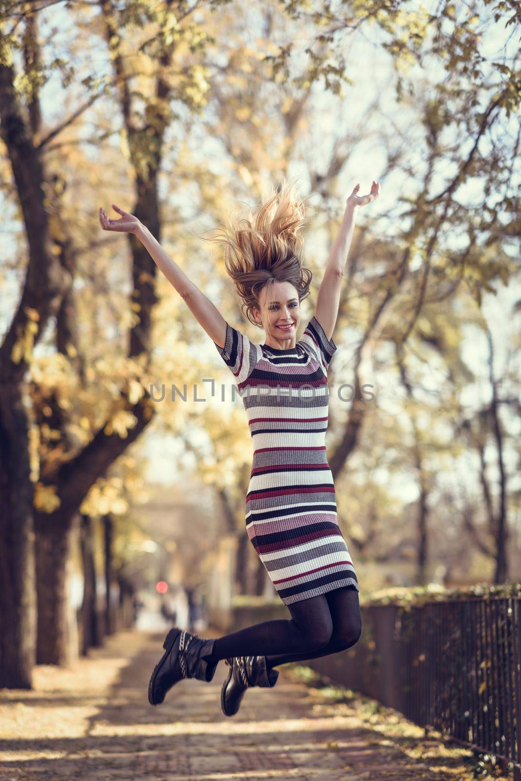 Young blonde woman jumping in the street. Beautiful girl in urban background wearing striped dress and black tights. Female with straight hair.