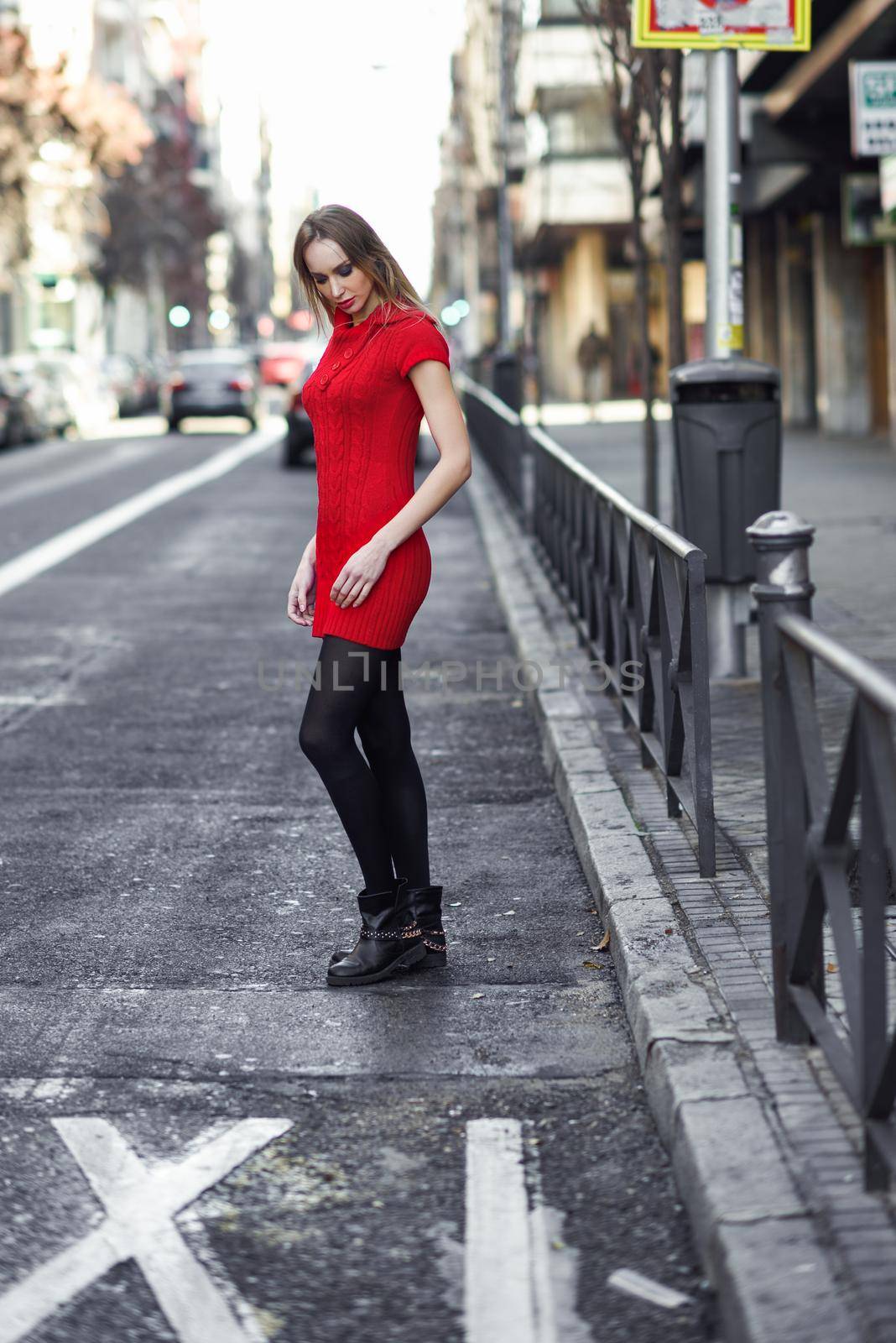 Young blonde woman standing in the street. Beautiful girl in urban background wearing red dress and black tights. Female with straight hair.