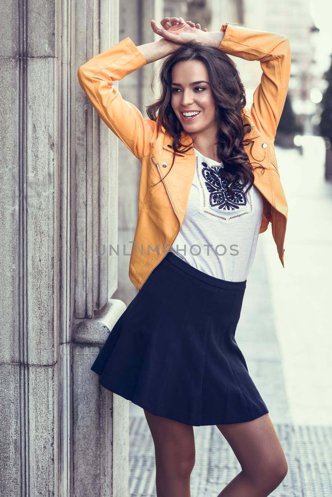 Young brunette woman, model of fashion, wearing orange modern jacket and blue skirt. Pretty caucasian girl with long wavy hairstyle smiling. Female raising her arms in urban background.