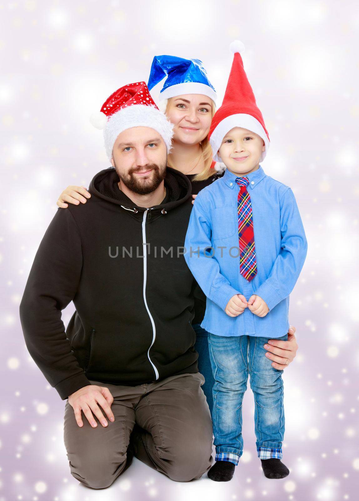Happy family of 3 people in caps of Santa Claus, the Christmas tree and the fireplace in the Christmas night.Blue Christmas festive background with white snowflakes.