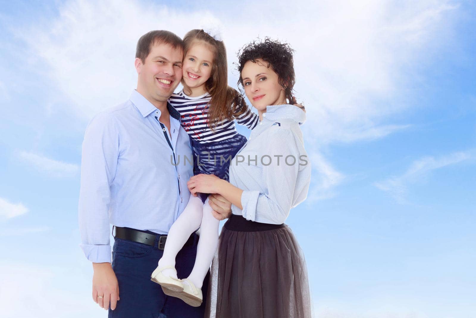 Happy young family, mom dad and little daughter.Parents keep the girl in her arms , and she hugs their neck.On the background of summer blue sky and fluffy clouds.
