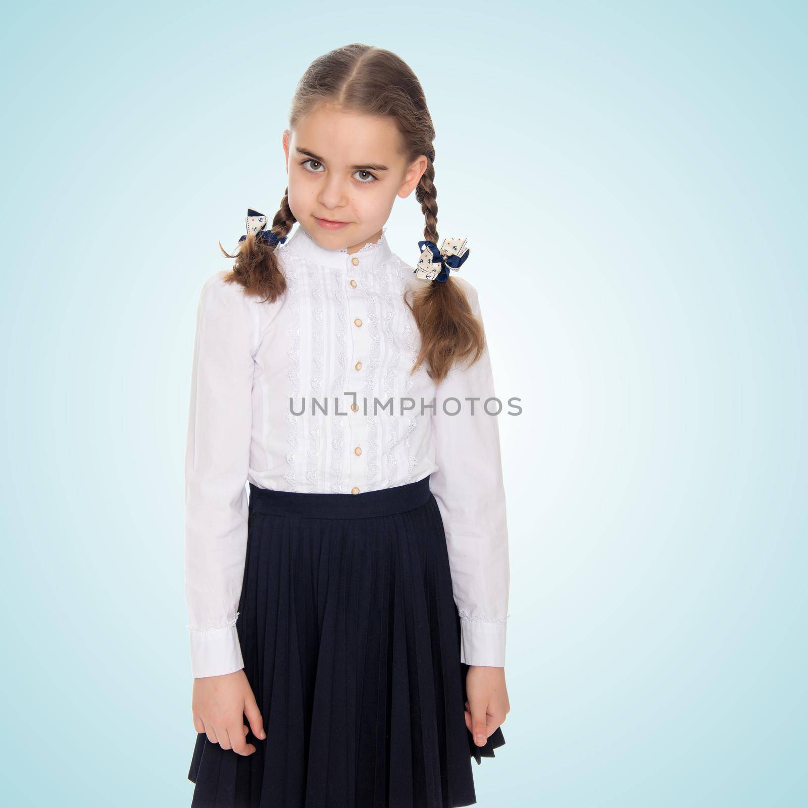 A beautiful little schoolgirl girl in a white blouse and black long skirt, with neatly braided pigtails on her head.She is standing right in front of the camera.Close-up.On the pale blue background.
