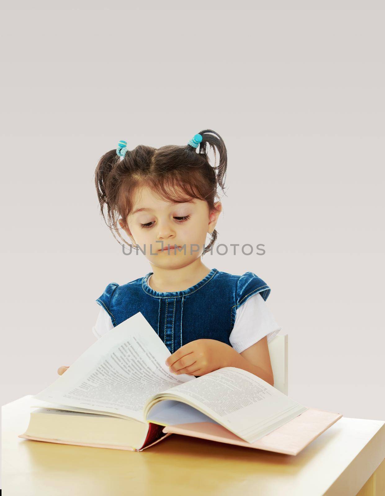 Pretty little girl in blue denim dress reading a book sitting at the table.On a gray background.