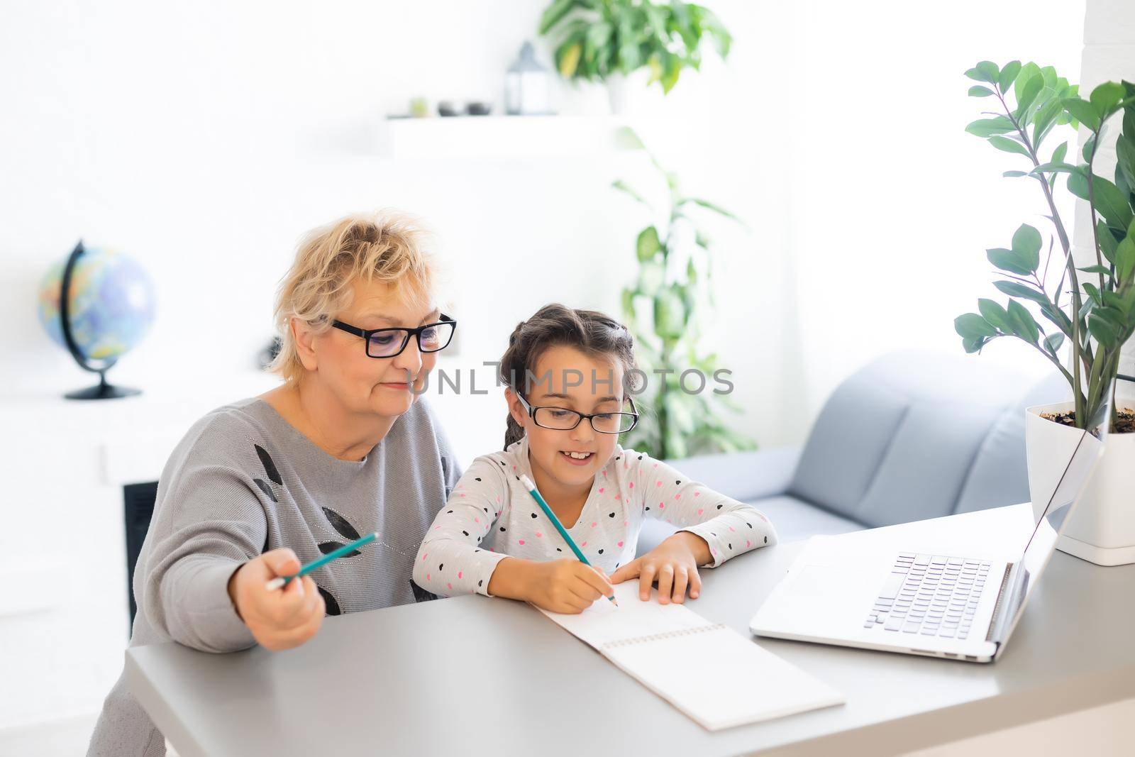 Mature grandmother helping child with homework at home. Satisfied old grandma helping her granddaughter studying in living room. Little girl writing on notebook with senior teacher sitting next to her