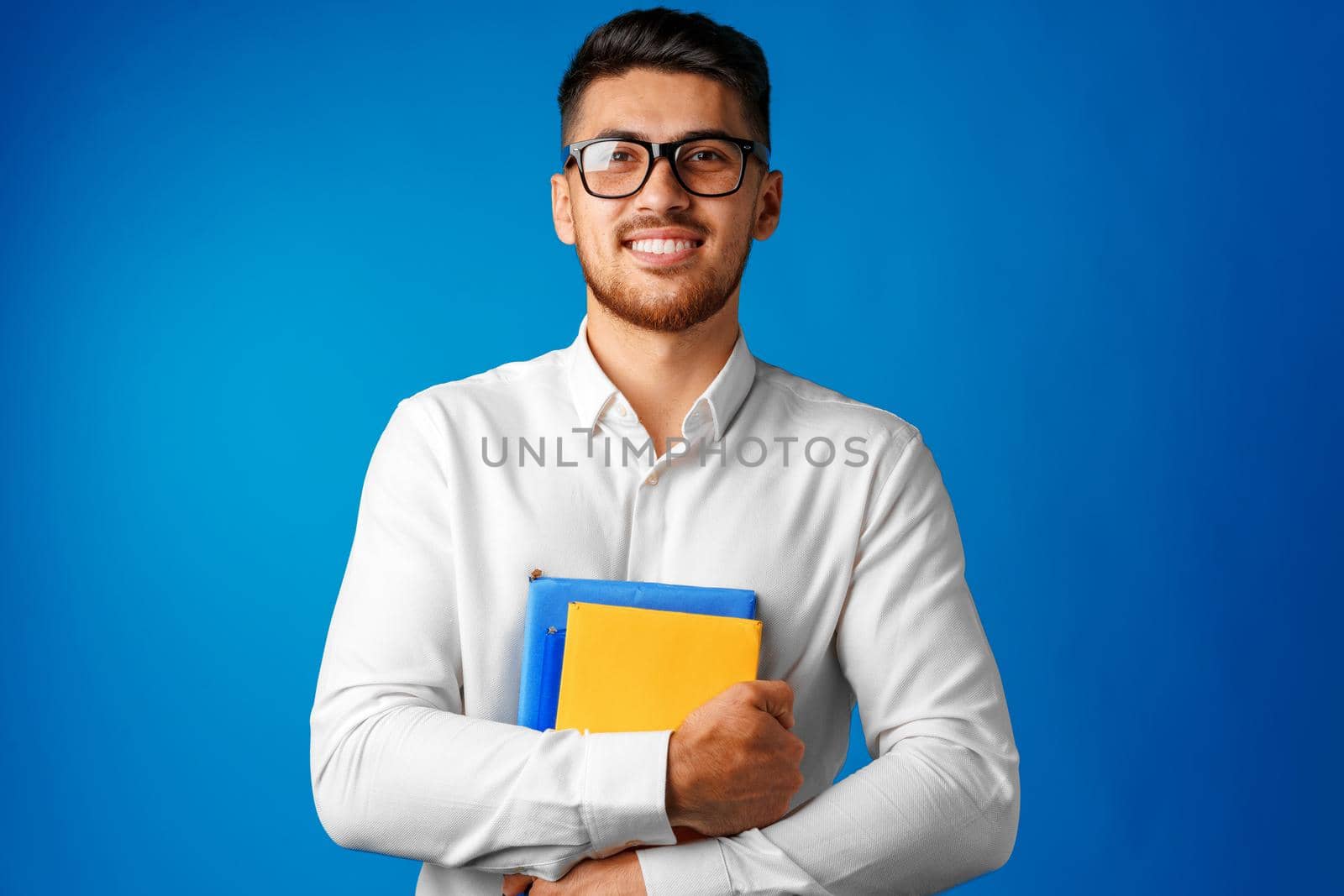 Man student in glasses holding books in arms against blue background close up