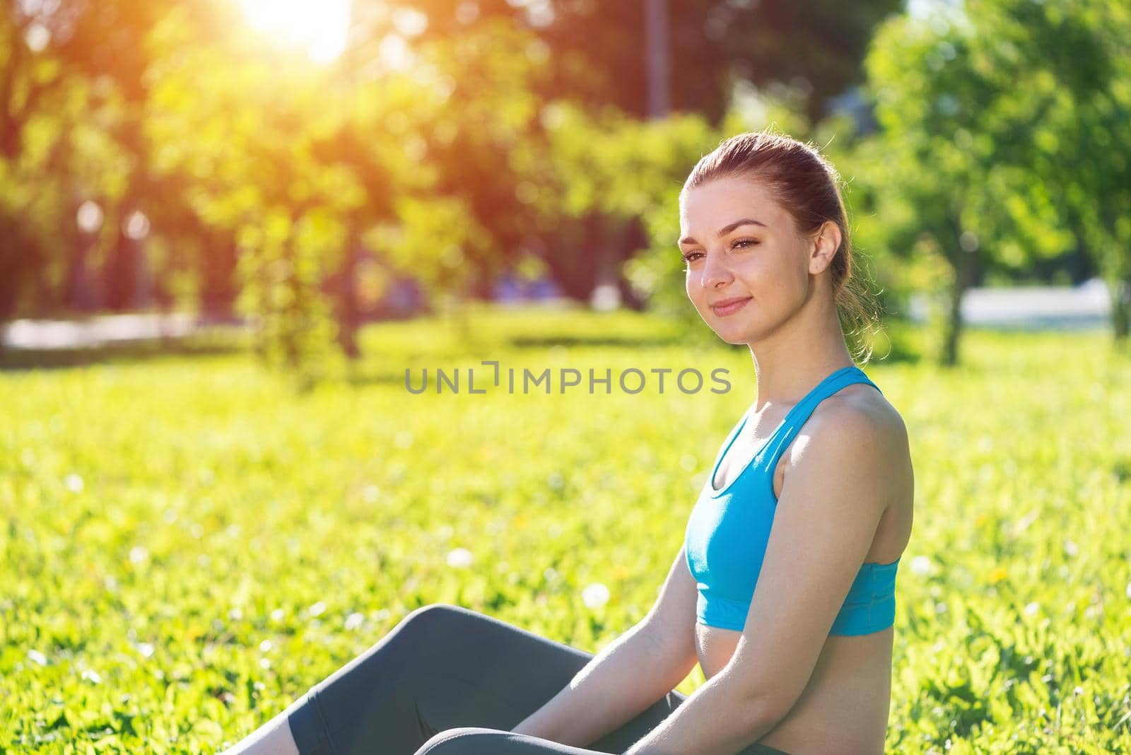 Beautiful smiling girl in activewear relax in park at sunrise. Portrait of young charming woman sitting on green grass. Training and meditation outdoor. Morning exercises and healthy lifestyle.