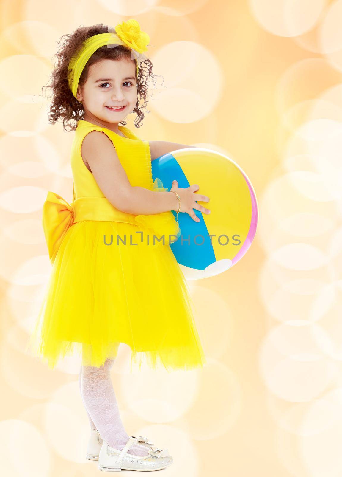 Laughing little girl in a bright yellow dress and bow on her head holding the ball. Girl posing sideways to the camera in full growth.Winter brown abstract background with white snowflakes.