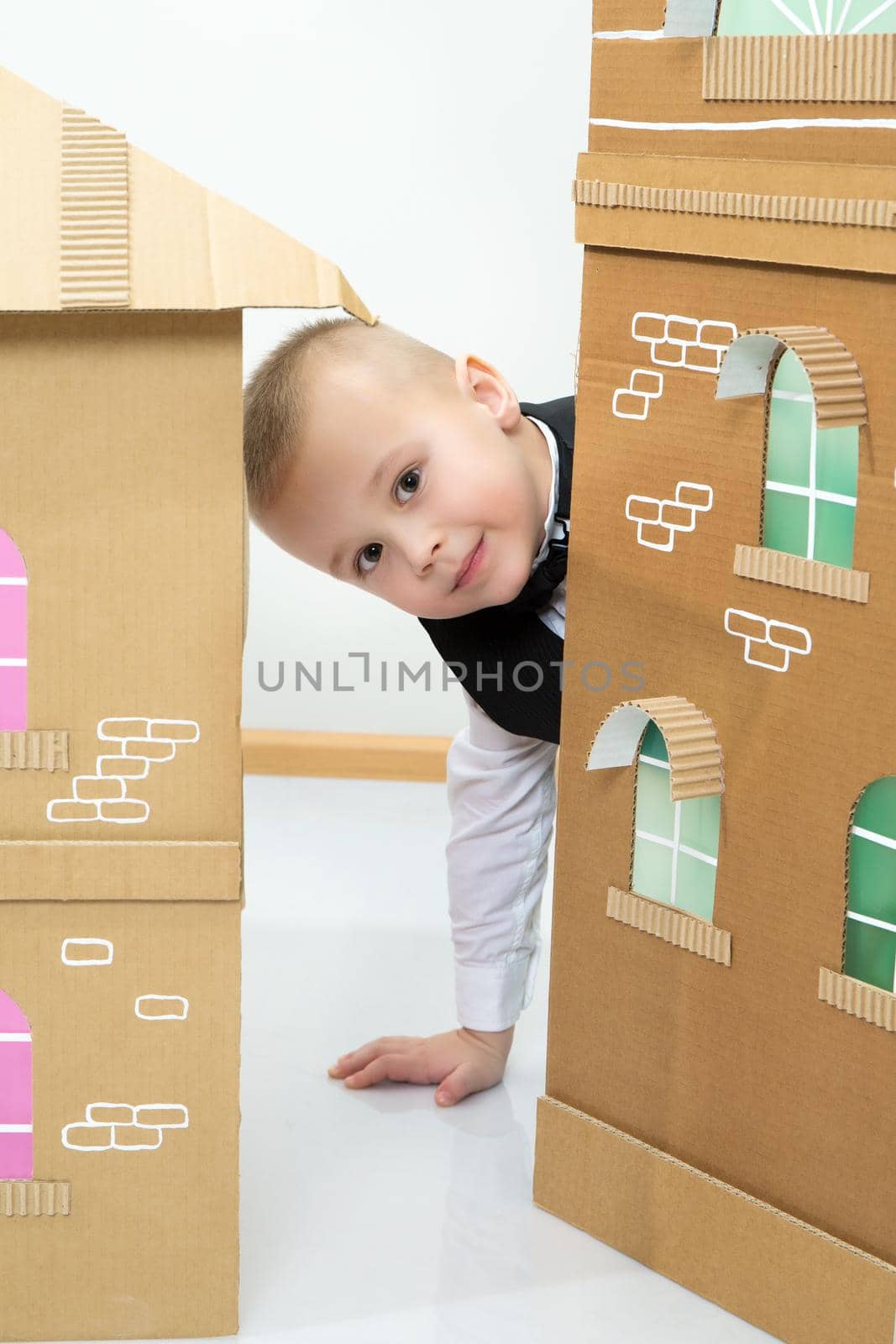 Beautiful little boy in a strict black suit , white shirt and tie.A boy peeks out from behind the cardboard house.