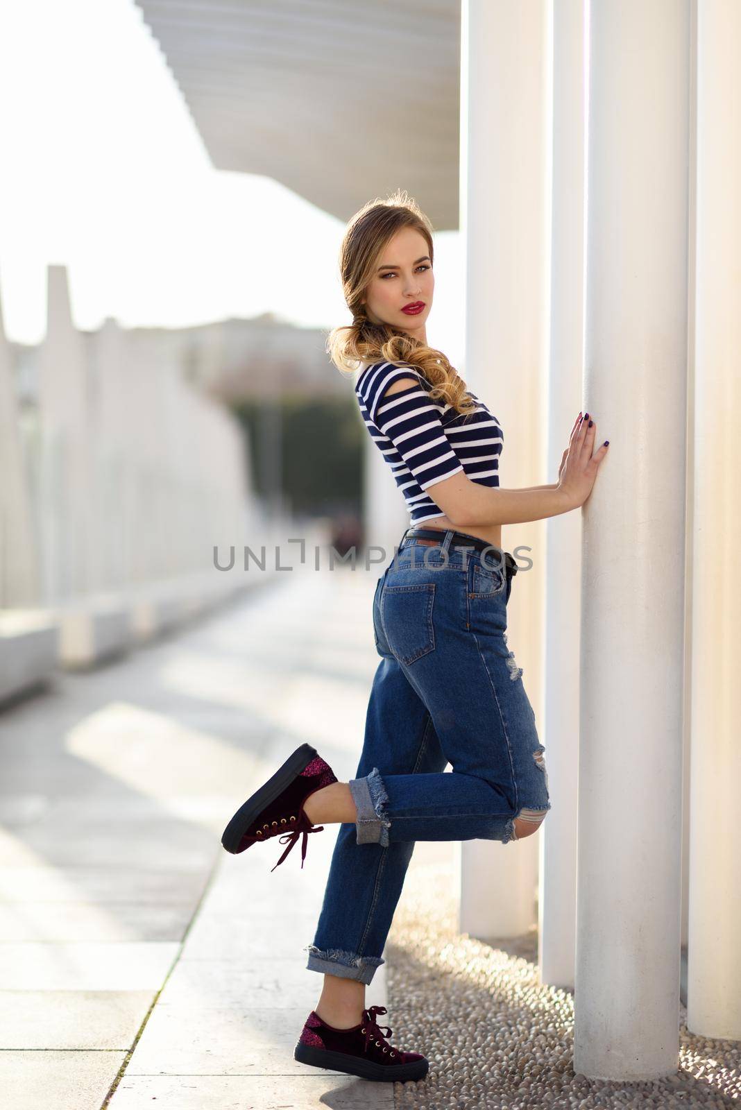 Blonde woman, model of fashion, possing in urban background. Beautiful young girl wearing striped t-shirt and blue jeans in the street. Pretty russian female with pigtail.