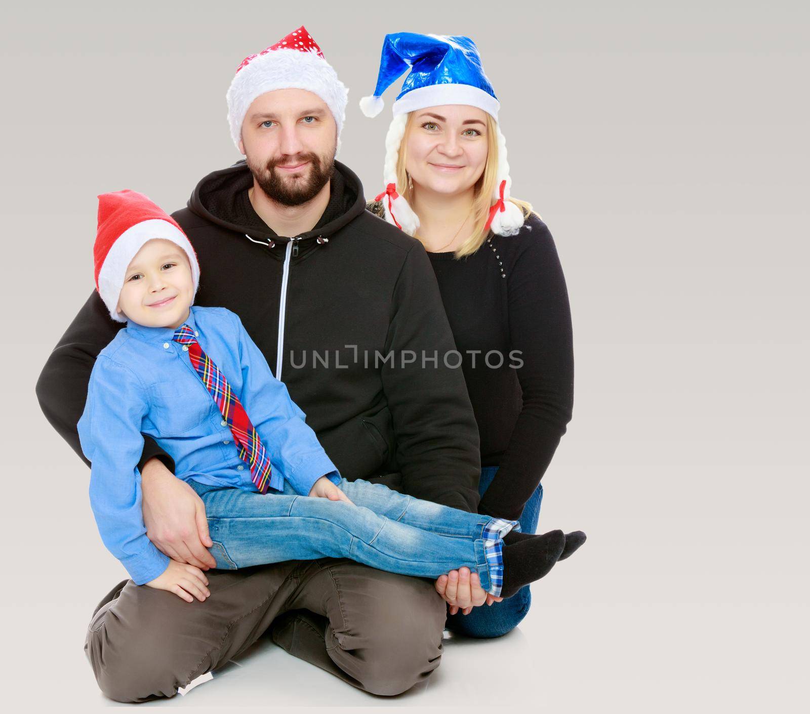 Happy family of 3 people in caps of Santa Claus, the Christmas tree and the fireplace in the Christmas night.On a gray background.