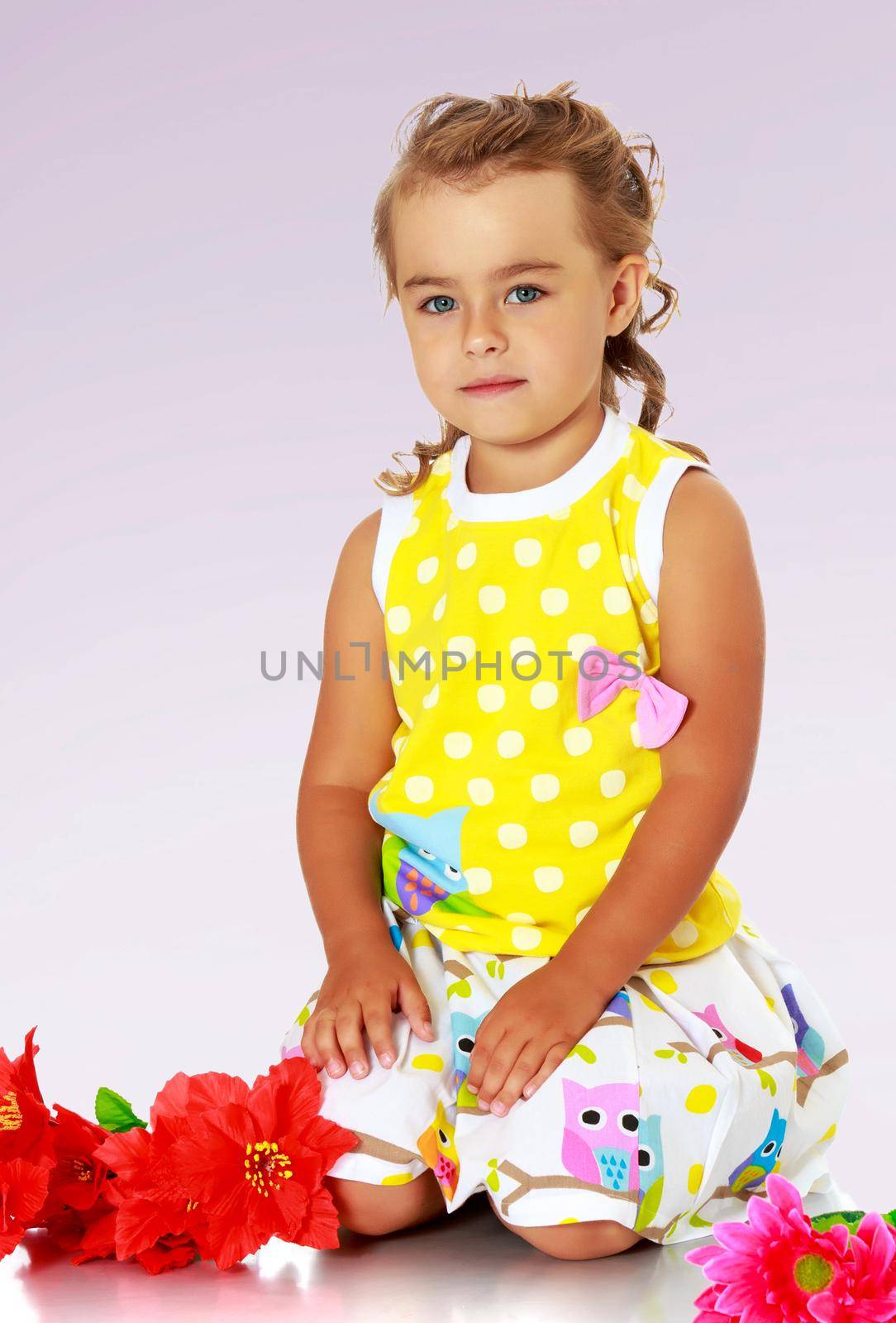 Beautiful little girl in a yellow summer dress kneeling on the floor. Beside her lay a red artificial flowers.Not a purple gradient background.