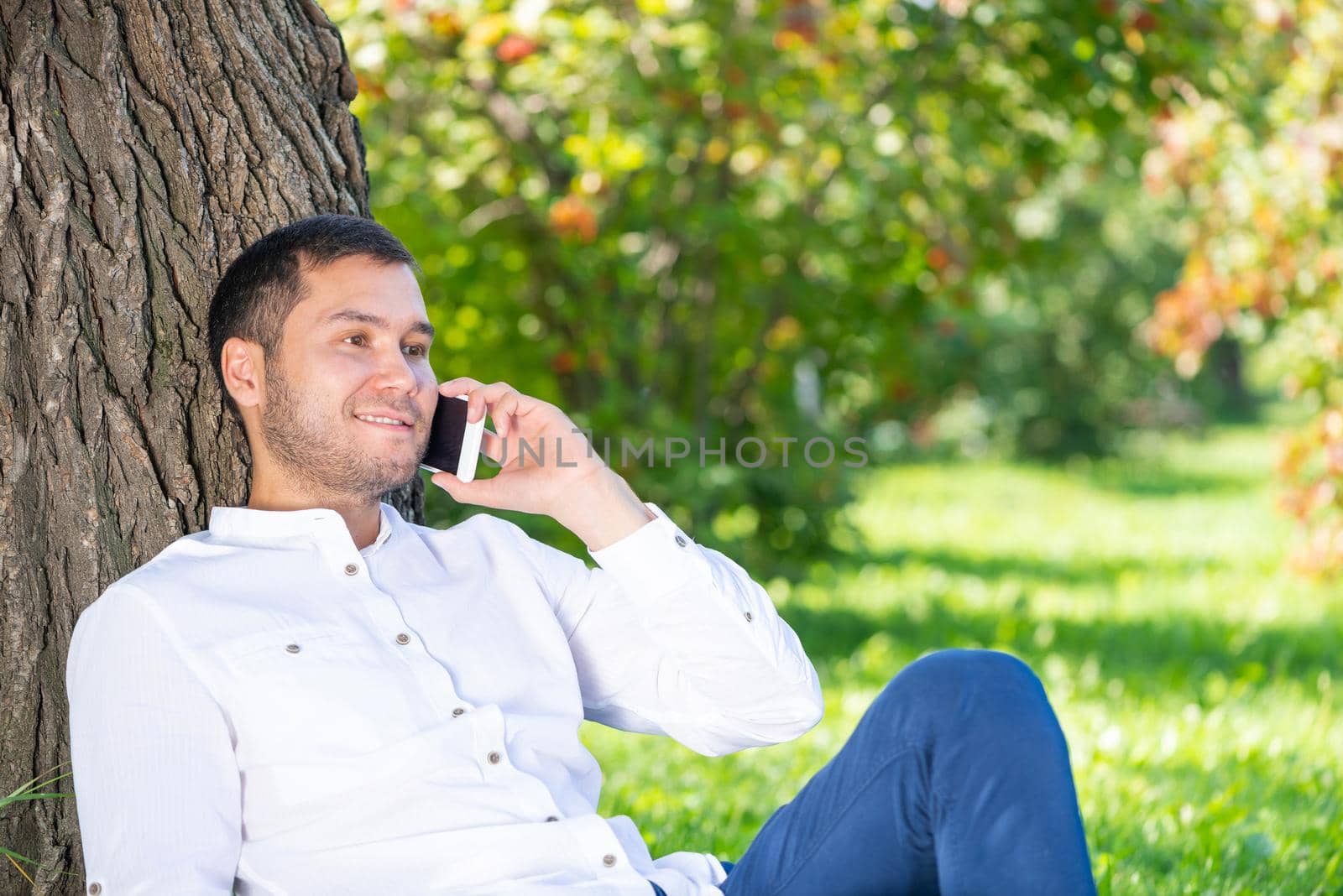 Young man talking on smartphone under tree in park on sunny day. Handsome businessman relaxing outdoors. Smiling man sitting on green grass leaning against tree. Mobile communication concept.
