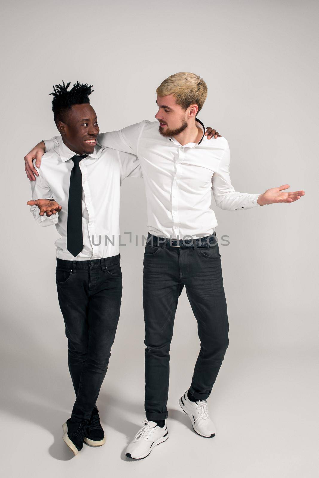 Joyful relaxed african and caucasian boys in white and black office clothes laughing and posing at white studio background with copy space. Full-length photo