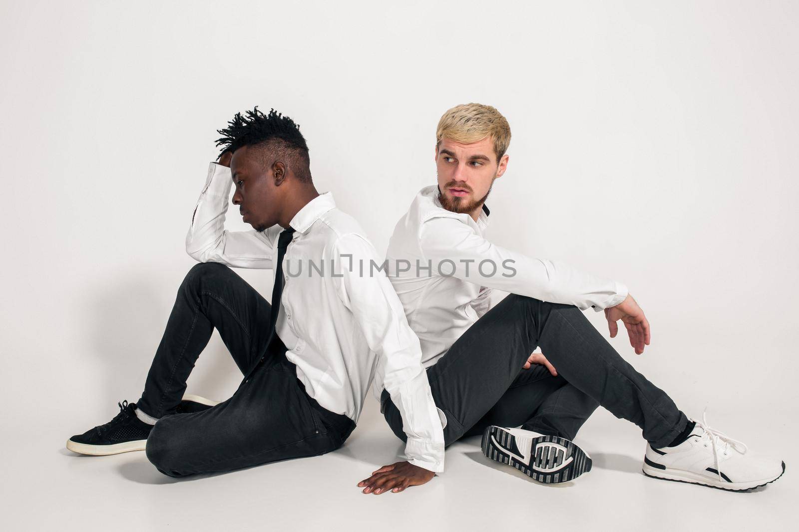 Friends. Two guys in white shirts and dark pants posing in the studio on a white background. Copy space. Two friends are sitting on the floor