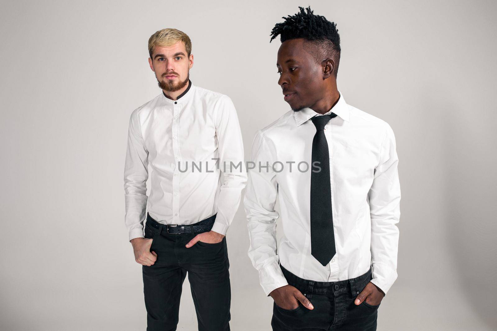 International friendship concept. Studio shot of two stylish young men: handsome bearded young man in white shirt and jeans standing next to his African-American friend on white background