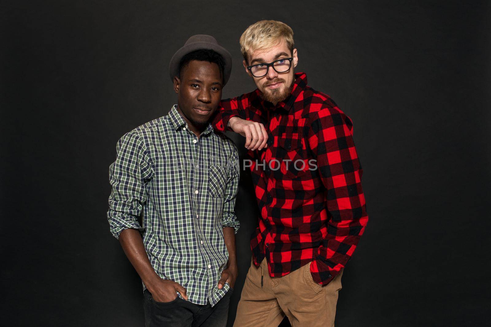 Studio shot of two stylish young men having fun. Handsome bearded hipster in a shirt in a cage standing next to his African-American friend in hat against a dark background. by nazarovsergey