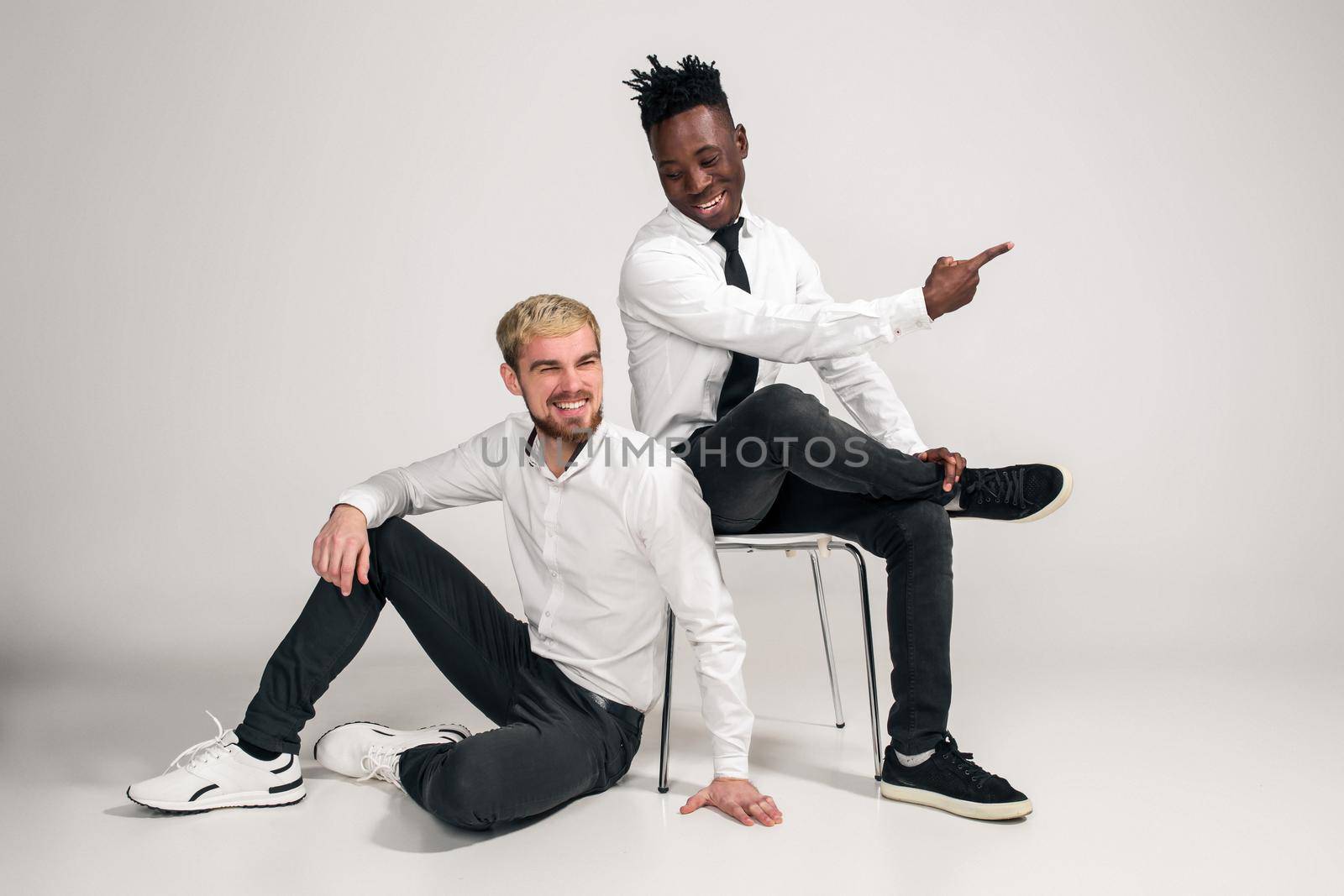 Joyful relaxed african and caucasian boys in white and black office clothes laughing and posing at white studio background with copy space. One man is sitting on the floor, the second is sitting on a chair