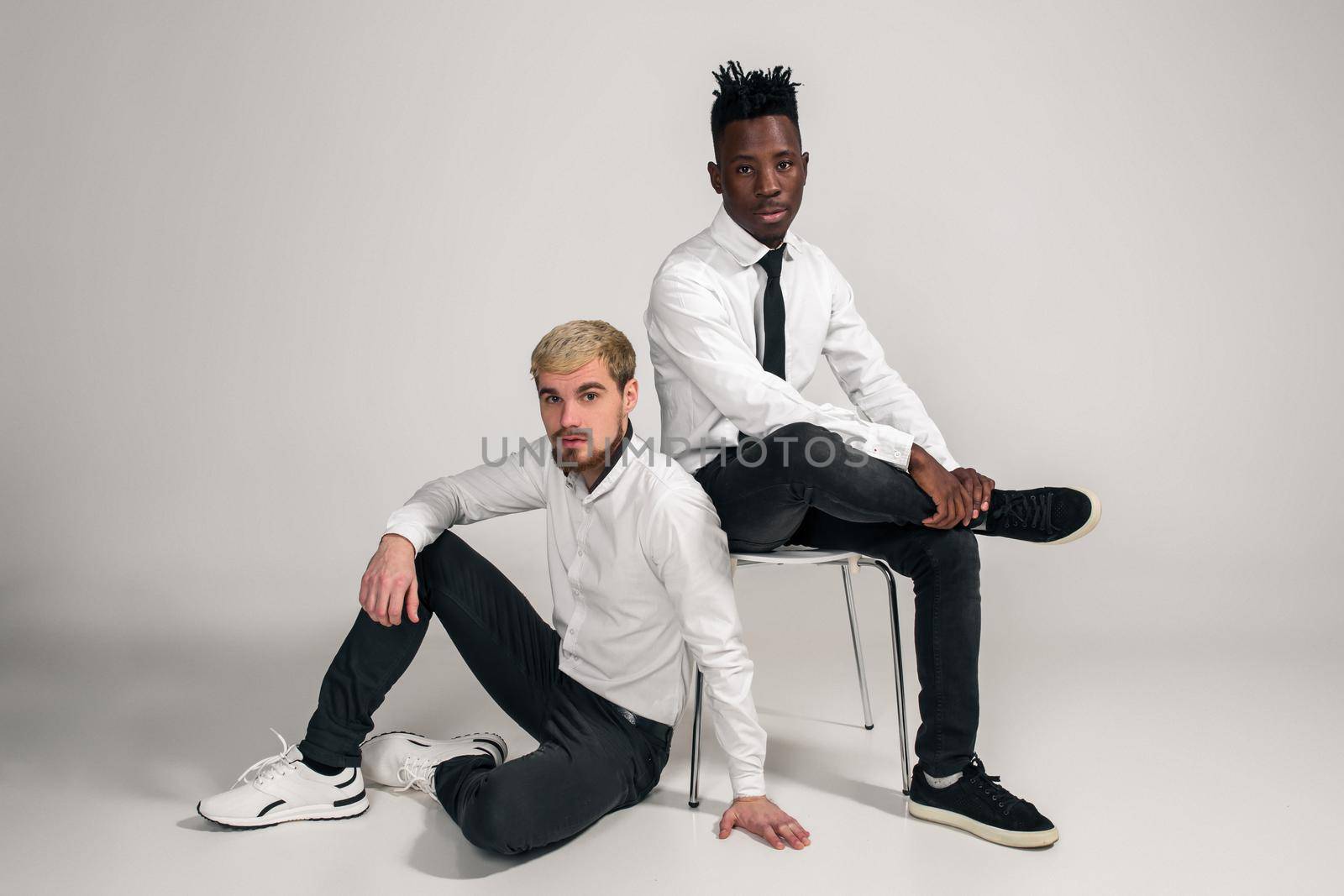 Joyful relaxed african and caucasian boys in white and black office clothes laughing and posing at white studio background with copy space. One man is sitting on the floor, the second is sitting on a chair