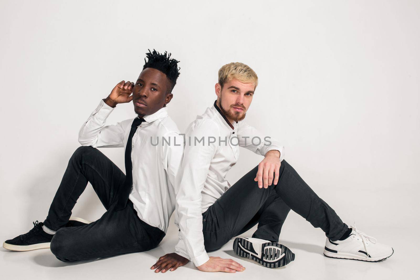 Friends. Two guys in white shirts and dark pants posing in the studio on a white background. Copy space. Two friends are sitting on the floor