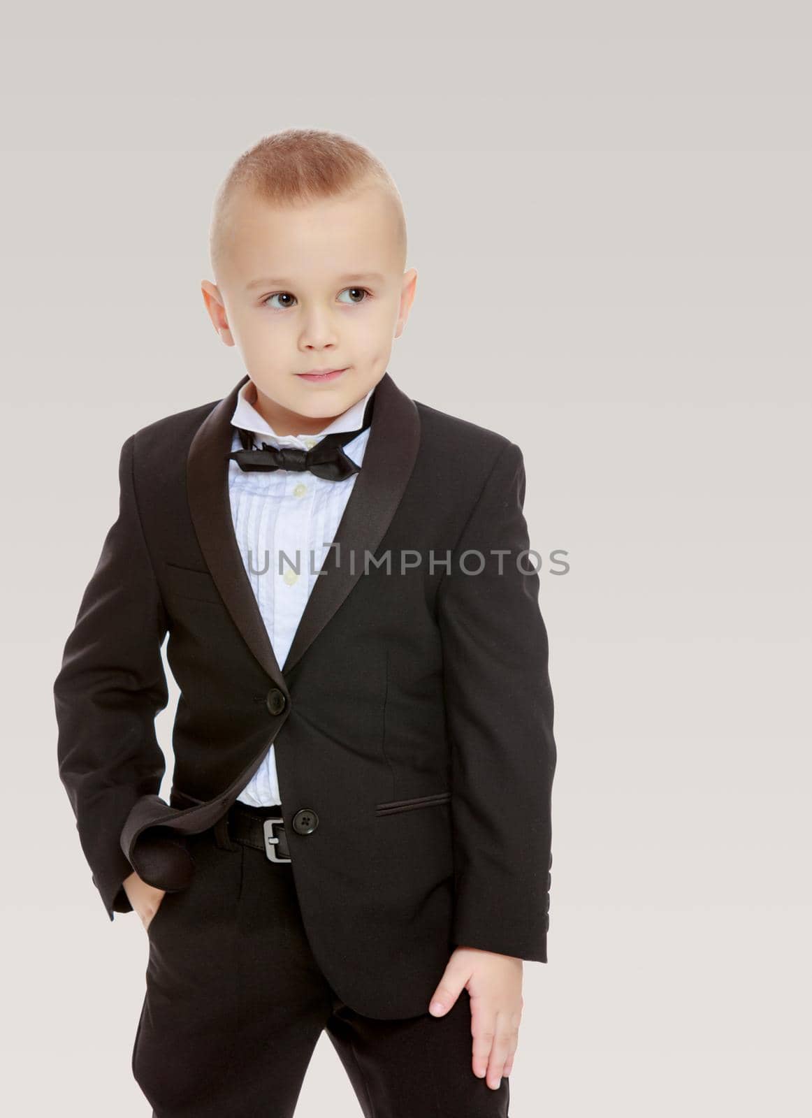 Beautiful little blond boy in a fashionable black suit with a tie.On a gray background.