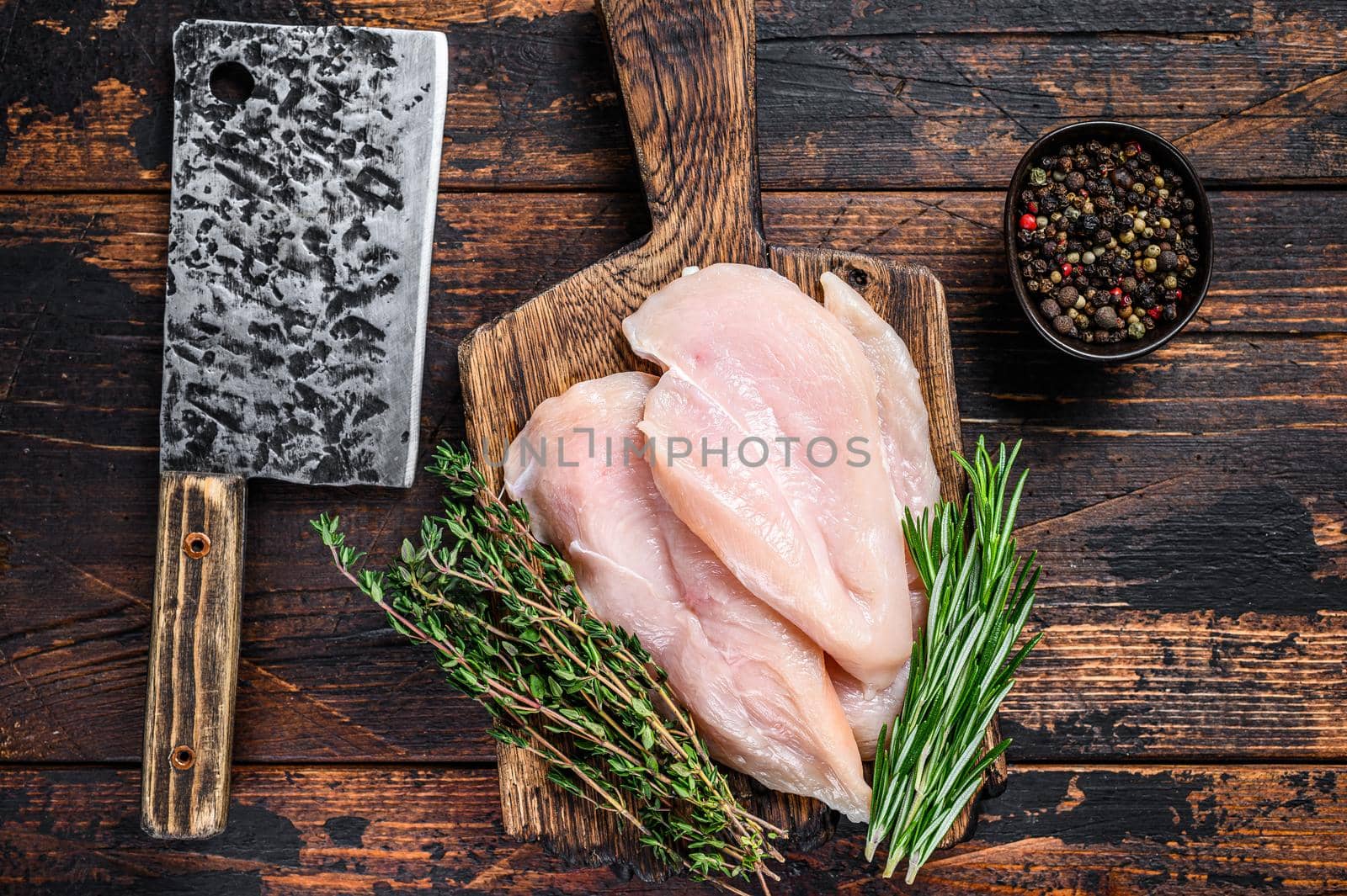 Raw sliced cut chicken breast fillet cutlets on a wooden cutting board with cleaver. Dark wooden background. Top view by Composter