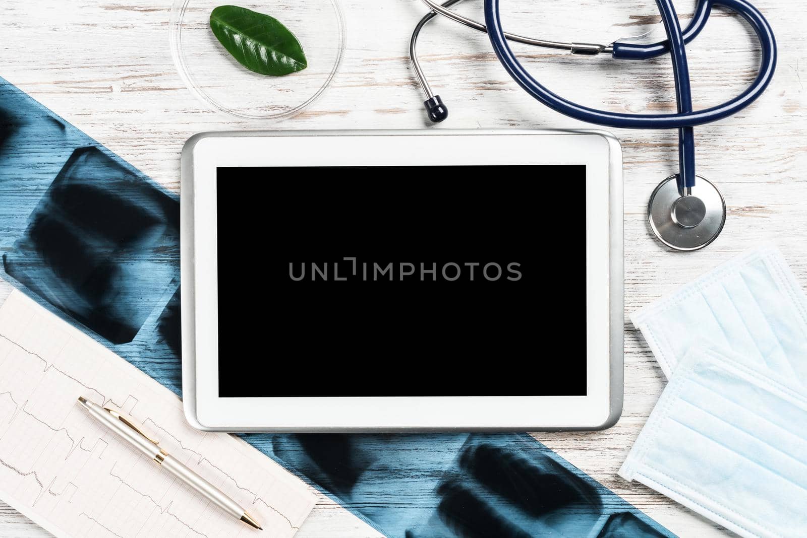 Medical diagnostics in modern hospital. Top view tablet computer with blank screen, x-ray images, stethoscope and cardiogram on wooden desk. Professional healthcare and digital technology in clinic.