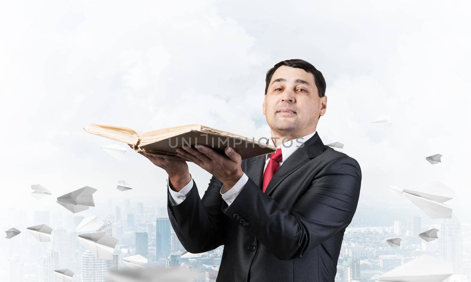 Serious businessman holding open book. Man in business suit standing on cloudy cityscape background with flying paper planes. Lawyer reading legal regulation book. Business accounting and consulting