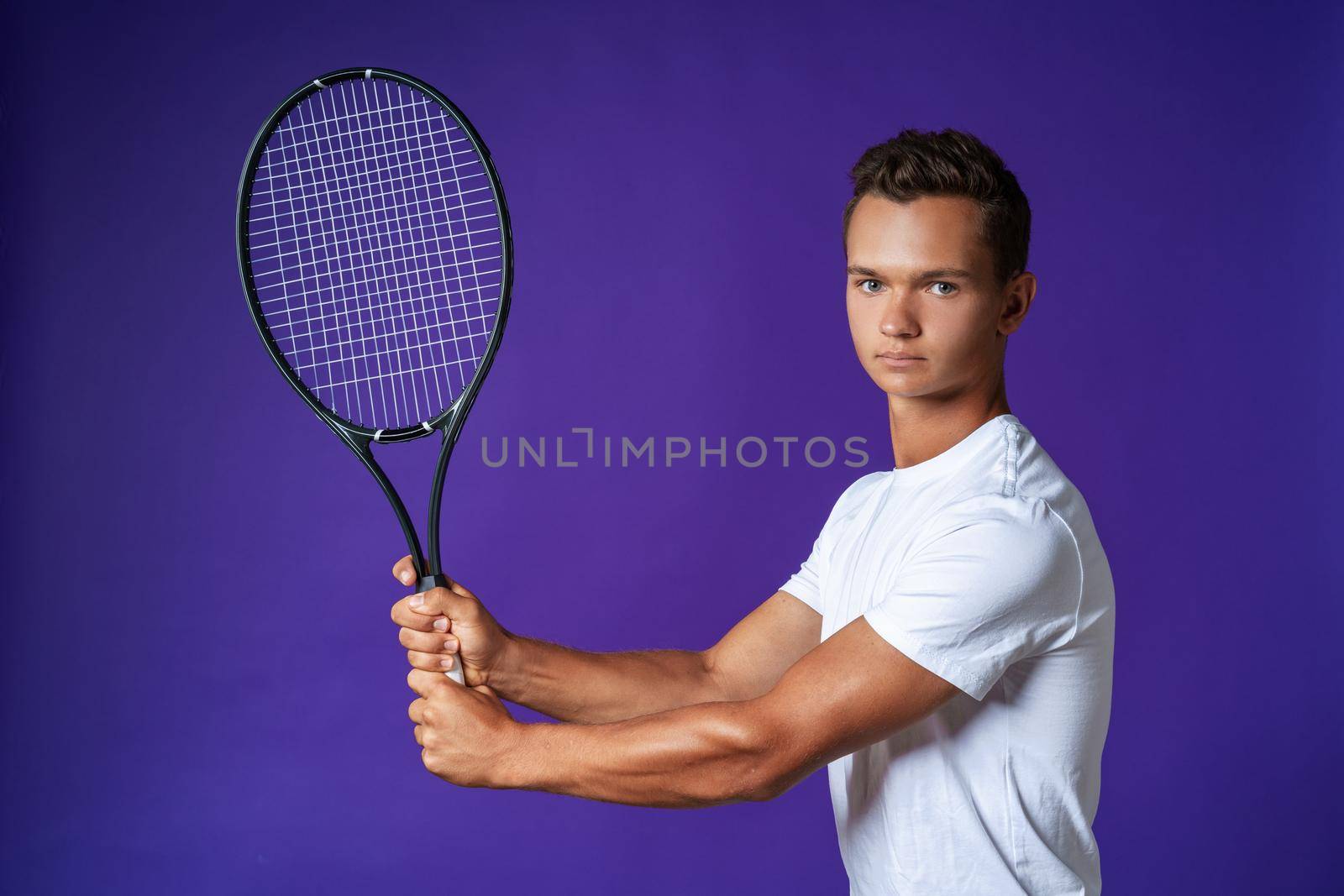 Caucasian young man tennis player posing with tennis racket against purple background close up