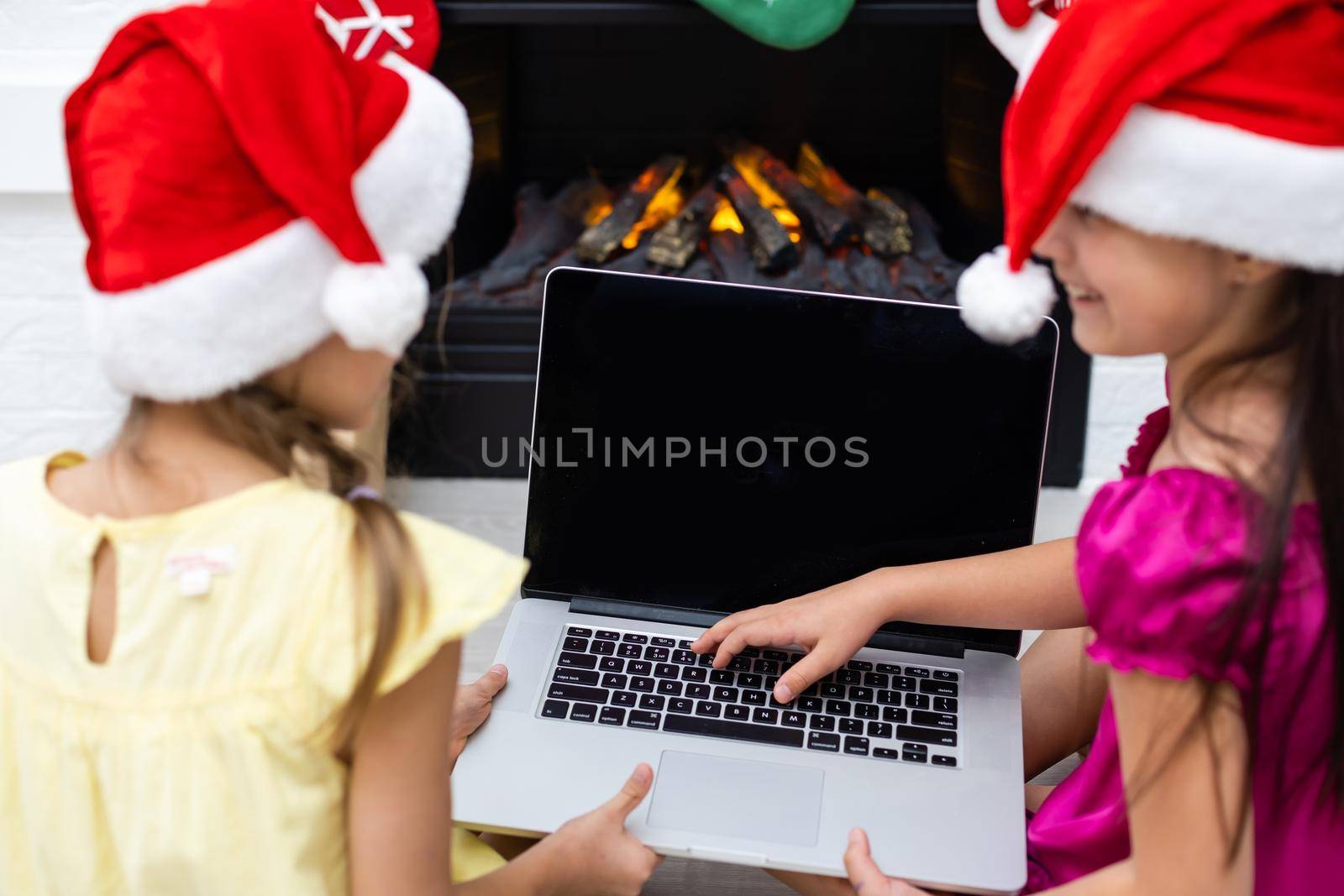 two little girls, sisters, girlfriends, play games, watch cartoons on a laptop on Christmas evening near the fireplace