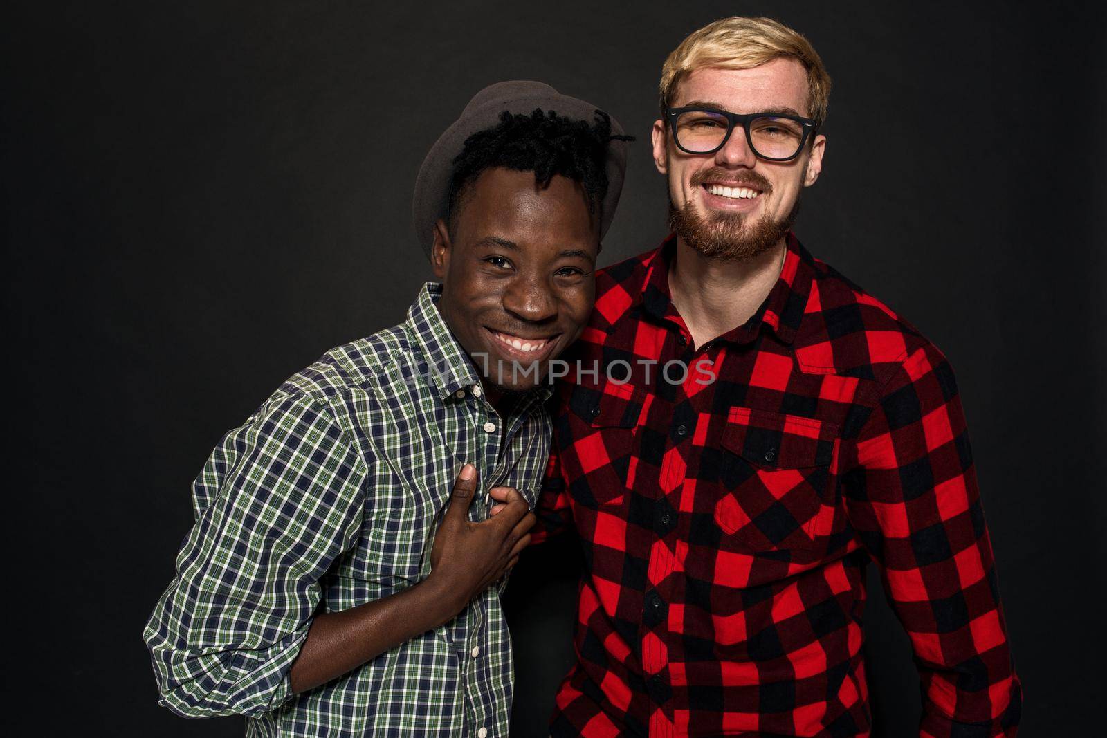 Studio shot of two stylish young men having fun. Handsome bearded hipster in a shirt in a cage standing next to his African-American friend in hat against a dark background. International friendship concept.