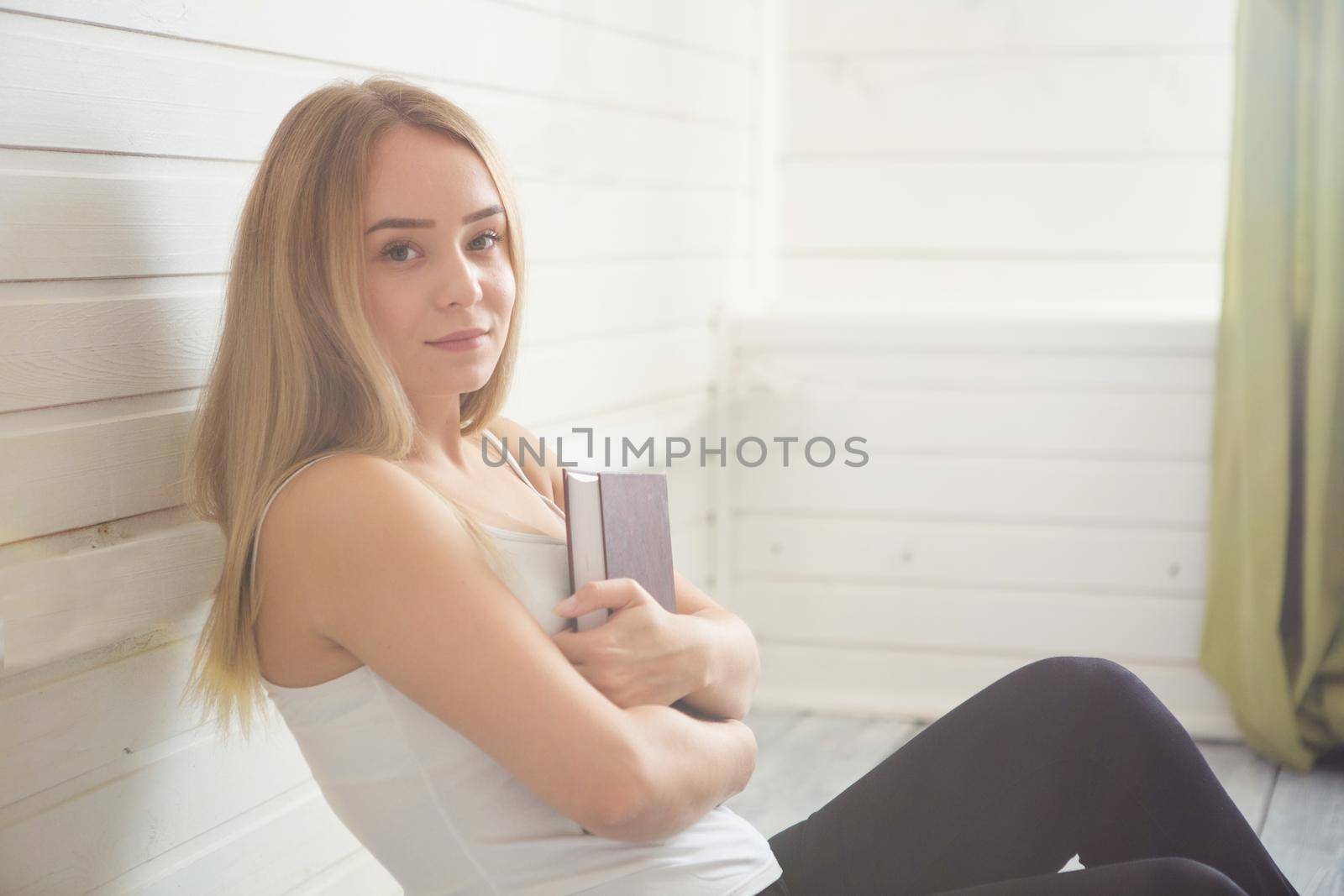 Lovely woman is sitting on the wooden floor with book over window.