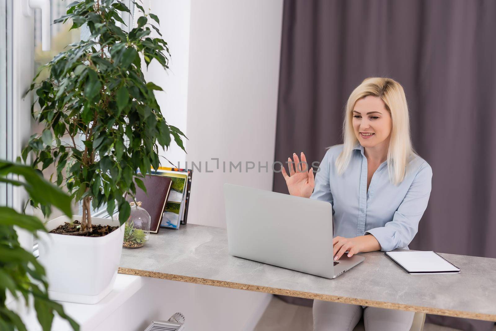 Young woman sitting at her desk in front of a gray laptop while daydreaming of nice memories or future in a modern bright office with large window