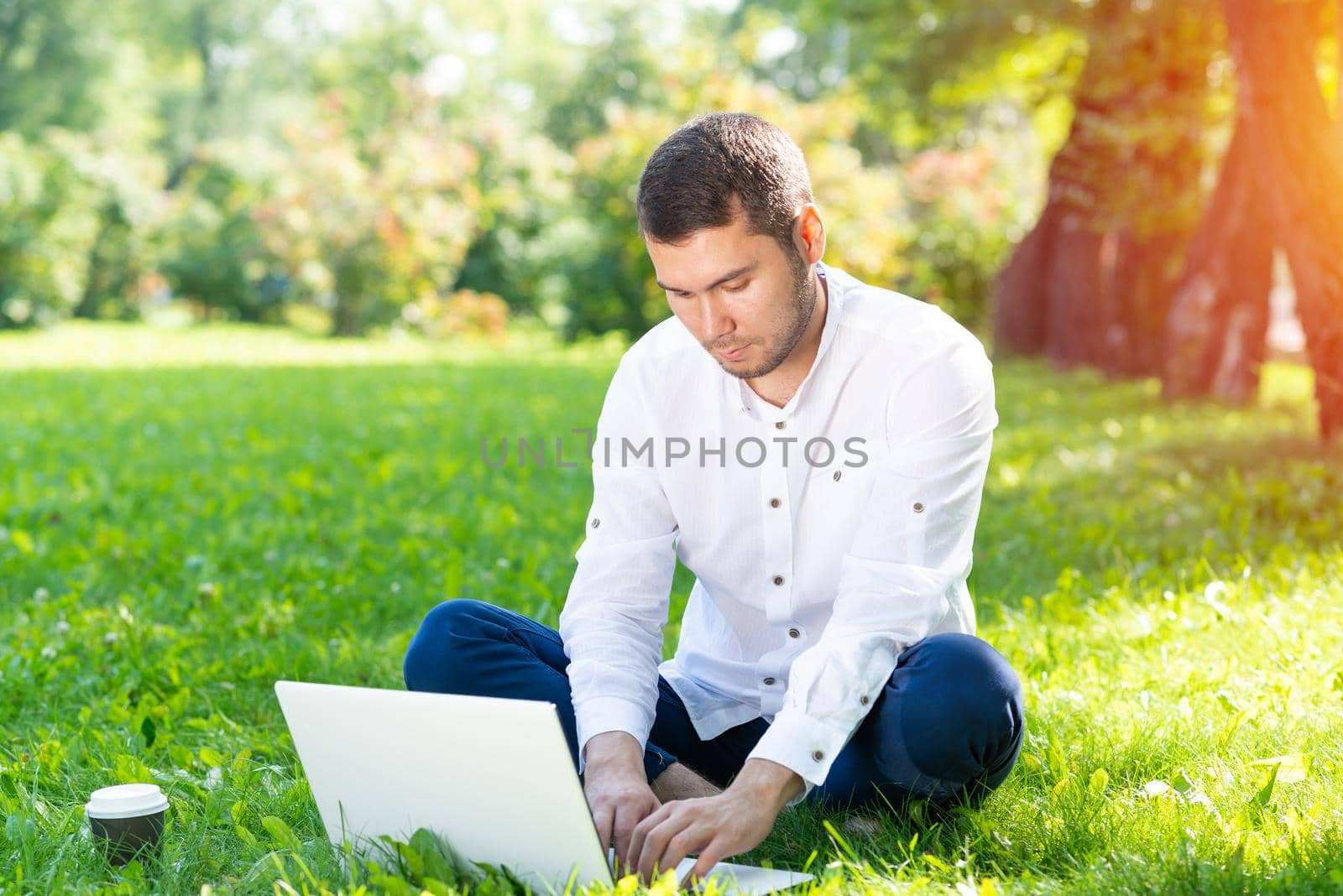 Young businessman using laptop computer on green grass in park. Handsome man in casual wear working with computer outdoors at sunny summer day. Freelance, blogging or remote work concept.