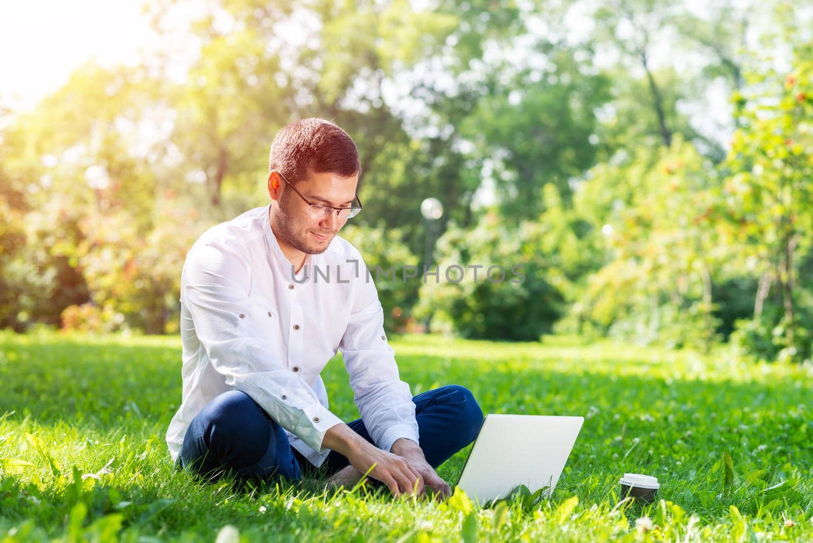 Young businessman sitting on green grass and using laptop computer. Handsome man working with computer in park at sunny summer day. Outdoors nature journey and relaxation. Freelance work concept.