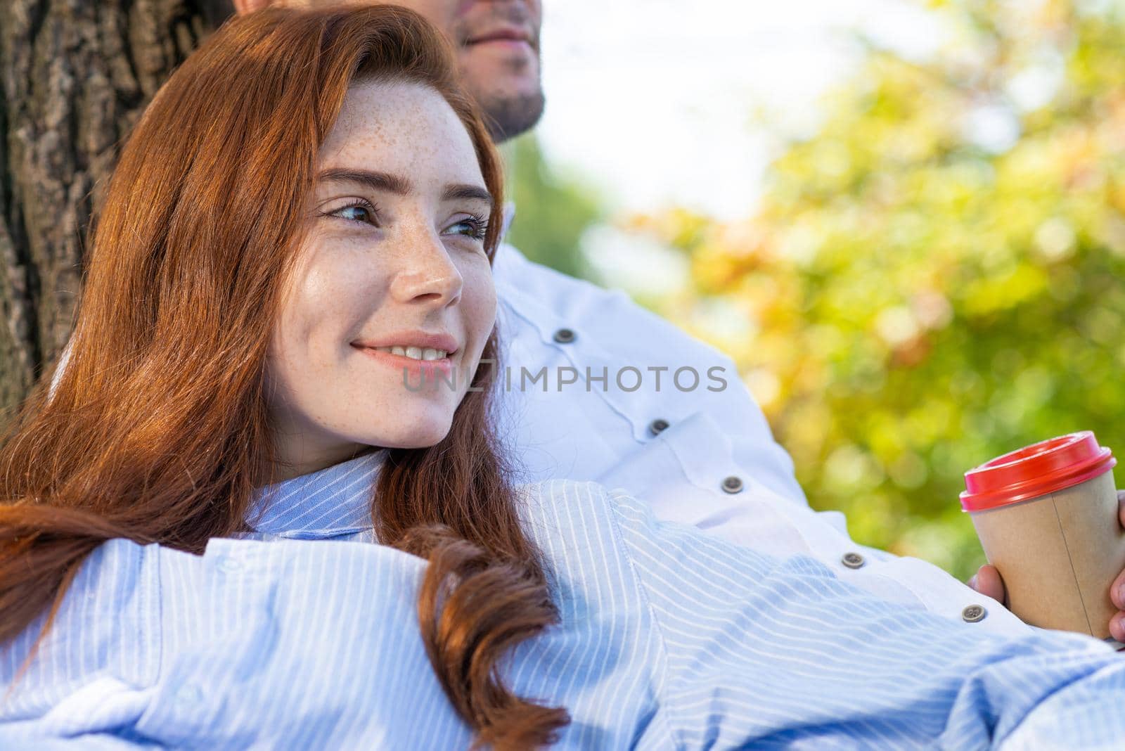 Young couple relaxing under tree in summer park on sunny day. Happy couple in love spend time outdoors together. Handsome man and pretty redhead girl enjoying each other. Romantic relationships.