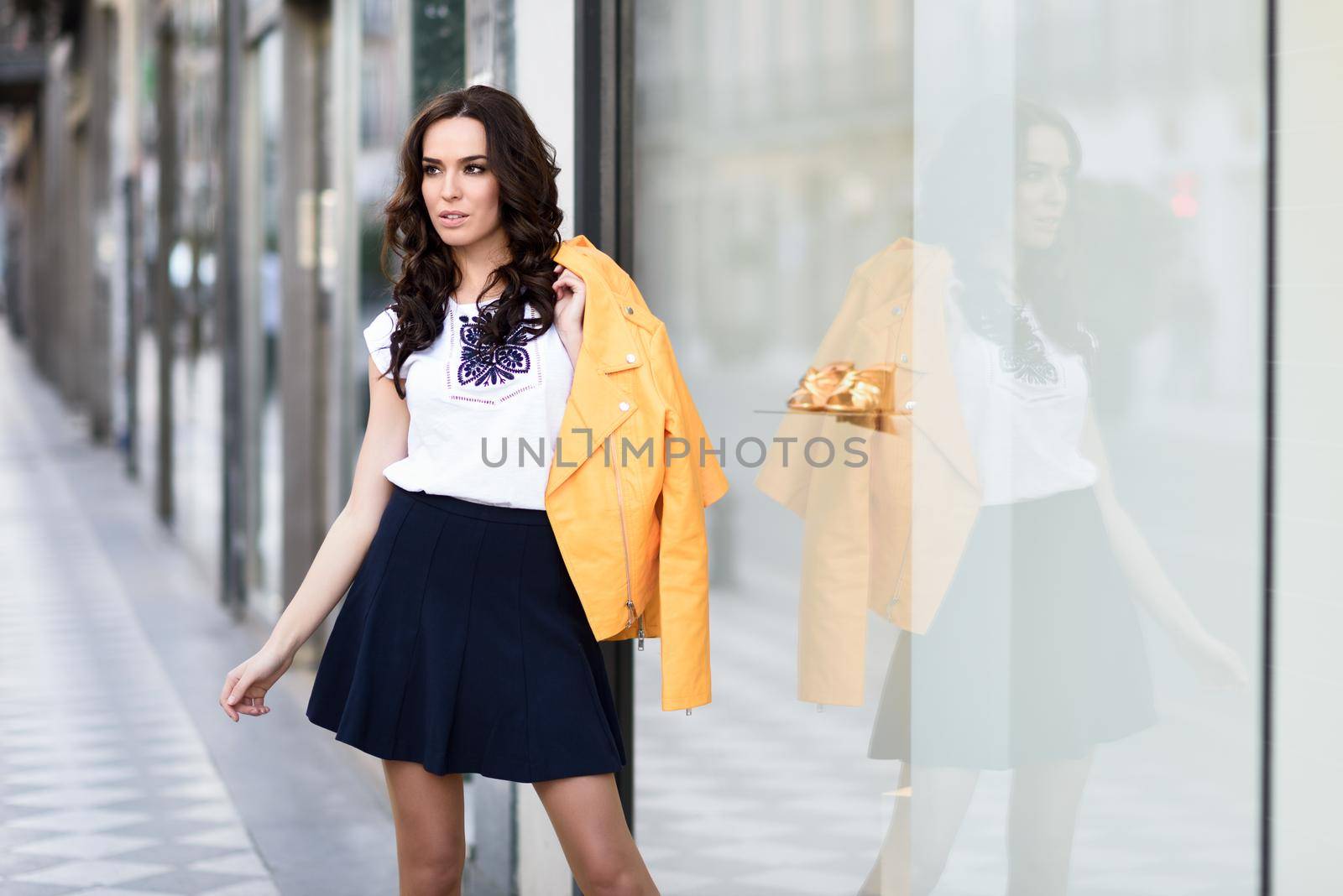 Young brunette woman, model of fashion, wearing orange modern jacket and blue skirt, standing in urban background.