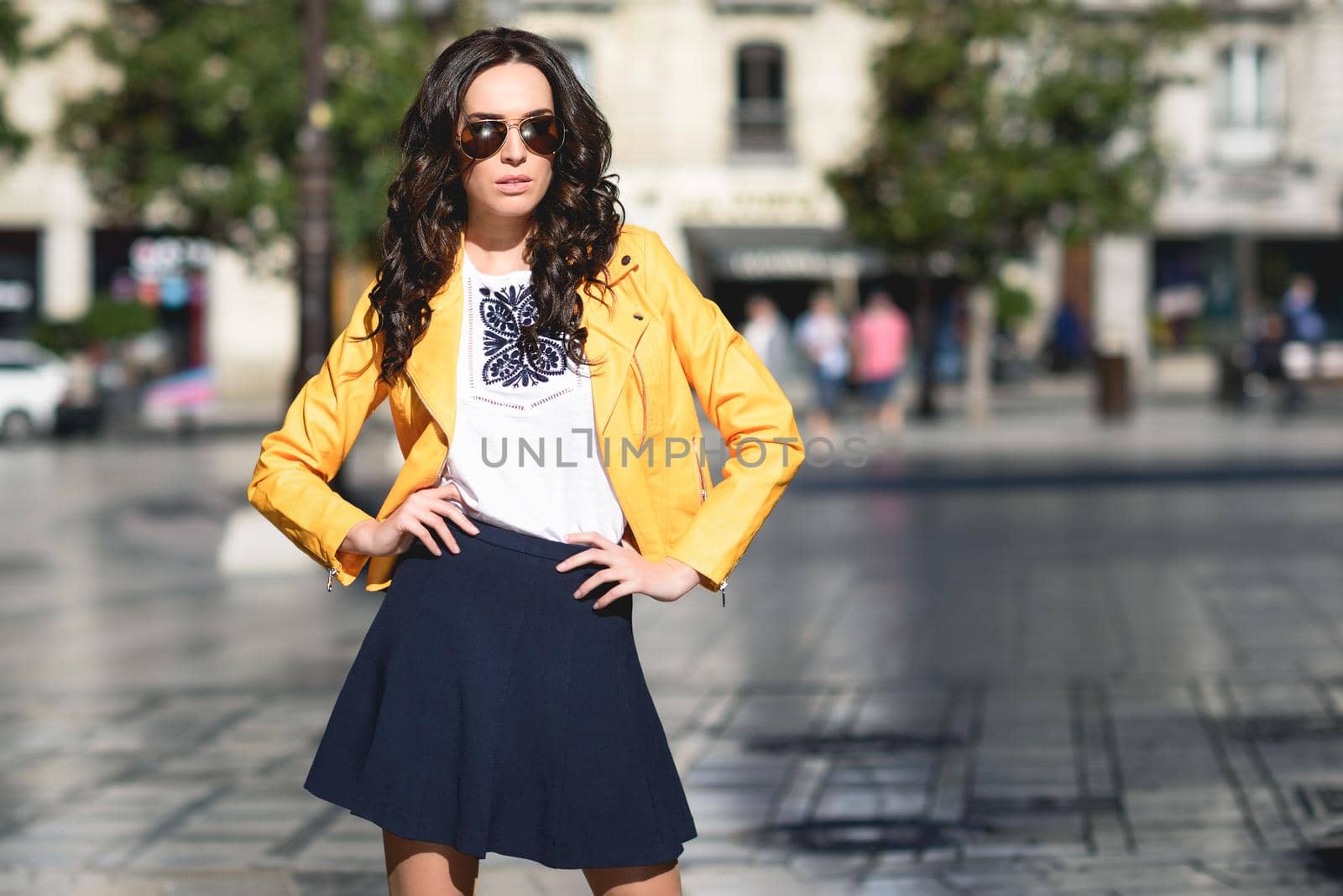 Young brunette woman with sunglasses. Girl, model of fashion, wearing orange modern jacket and blue skirt, standing in urban background.