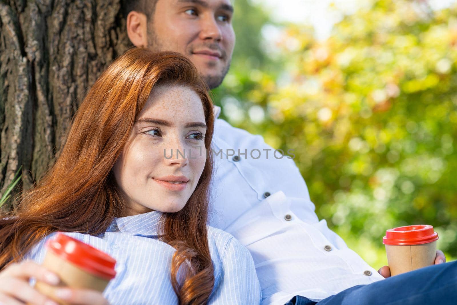 Young couple relaxing under tree in summer park on sunny day. Happy couple in love spend time outdoors together. Handsome man and pretty redhead girl enjoying each other. Romantic relationships.