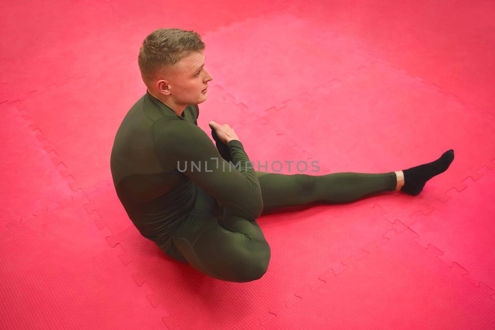 Young sports guy in a green thermal suit does exercises on the gym floor.
