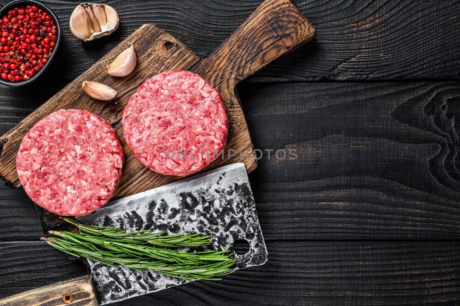 Raw steak cutlets with mince beef meat and rosemary on a wooden cutting board with meat cleaver. Black Wooden background. Top view. Copy space by Composter