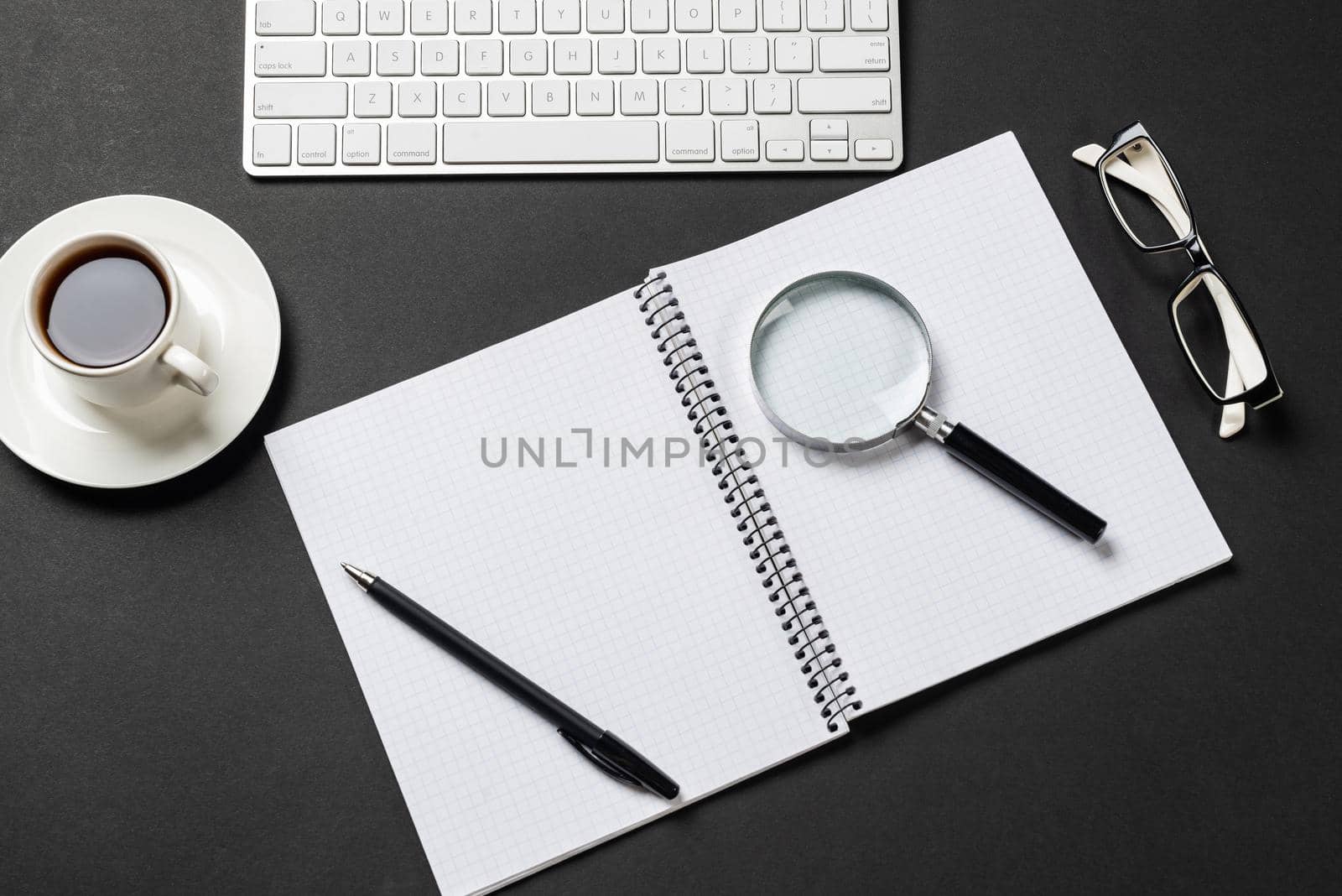Top view of modern workplace with magnifying glass. Flat lay black surface with wireless keyboard, cup of coffee and notebook. Top view home workspace and freelance. Consulting, audit and accounting.