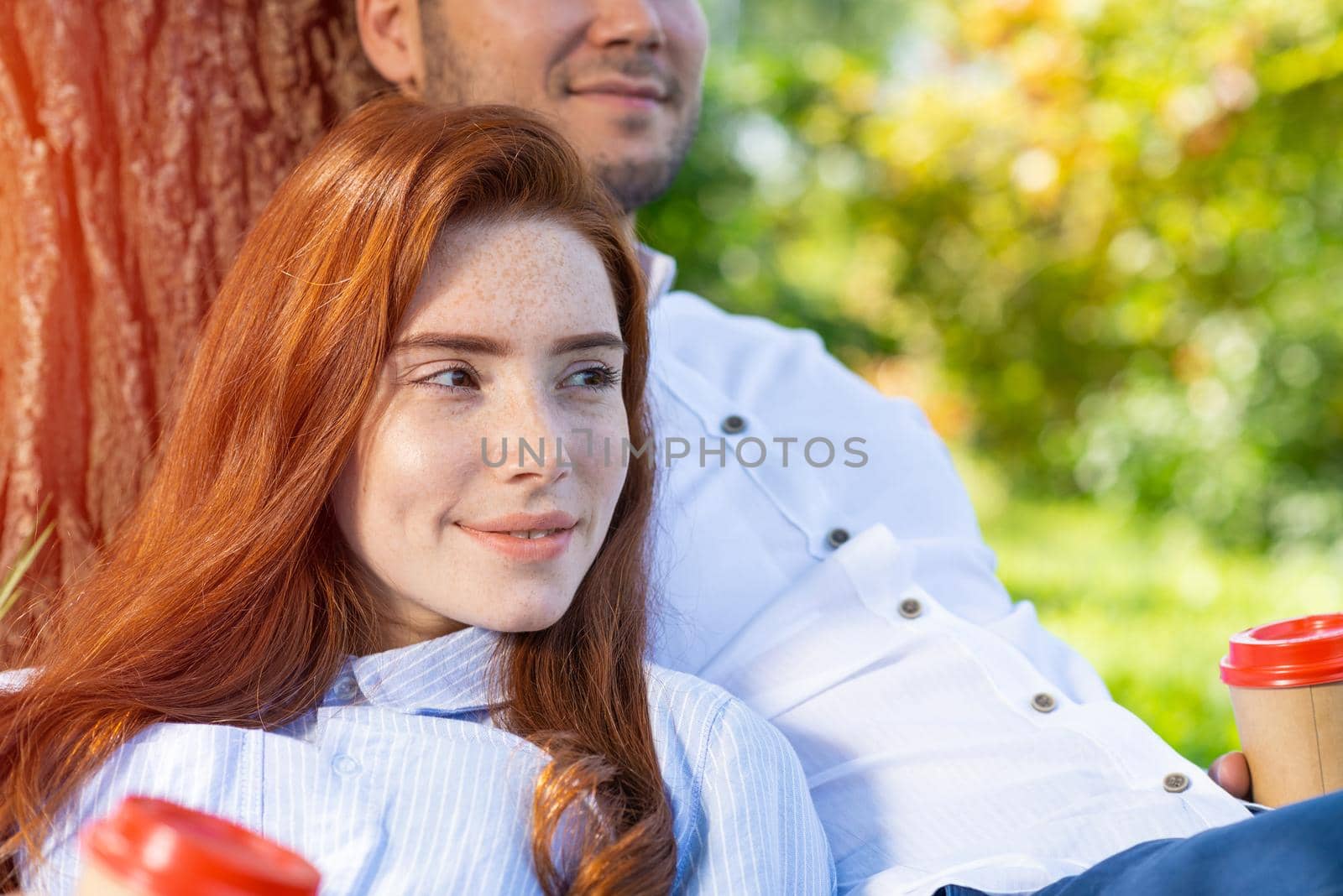Young couple relaxing with coffee under tree in park on sunny day. Happy couple in love spend time outdoors together. Handsome man and pretty redhead girl sitting on green grass leaning against tree