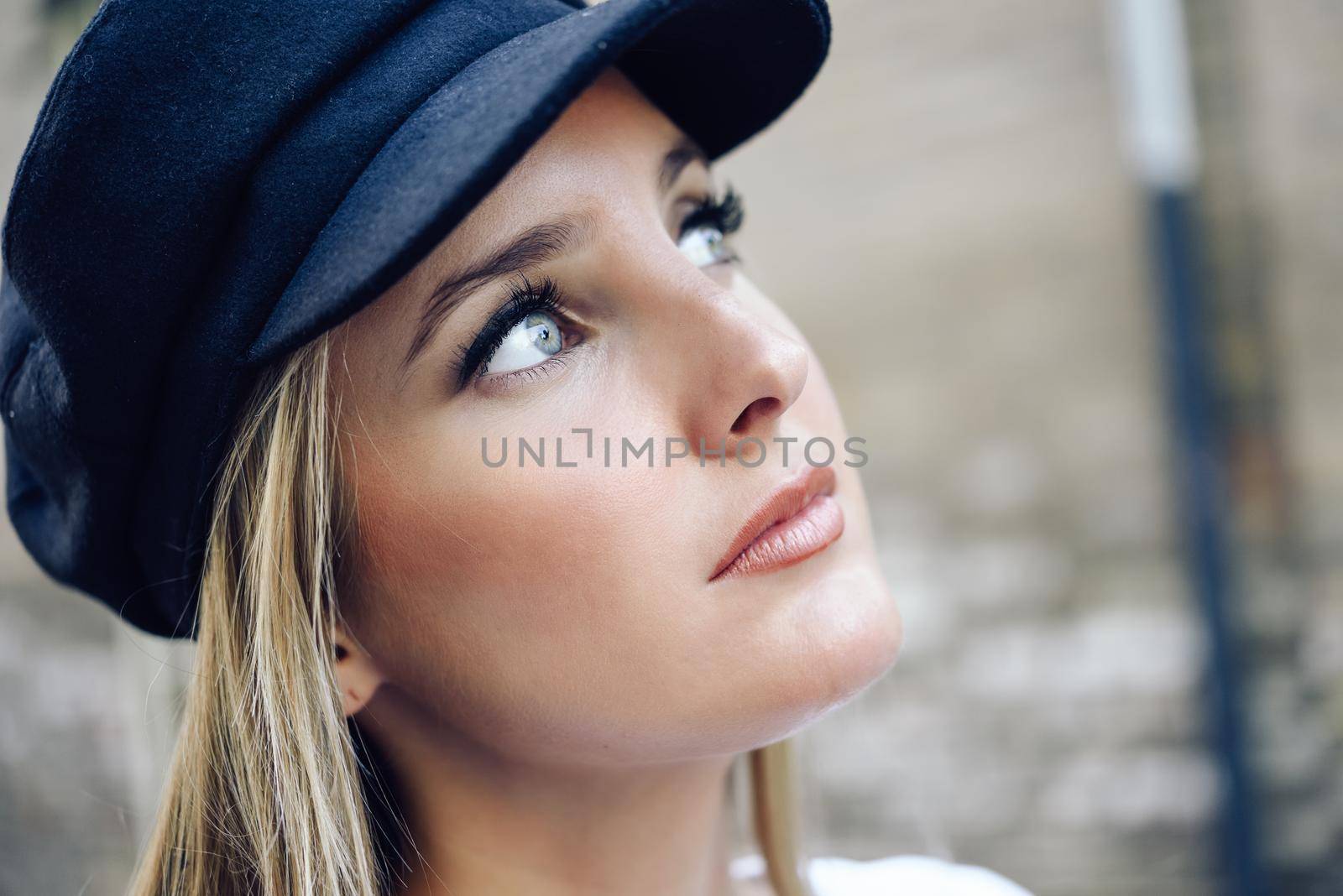 Young blonde woman in defocused urban background. Girl with blue eyes wearing cap.