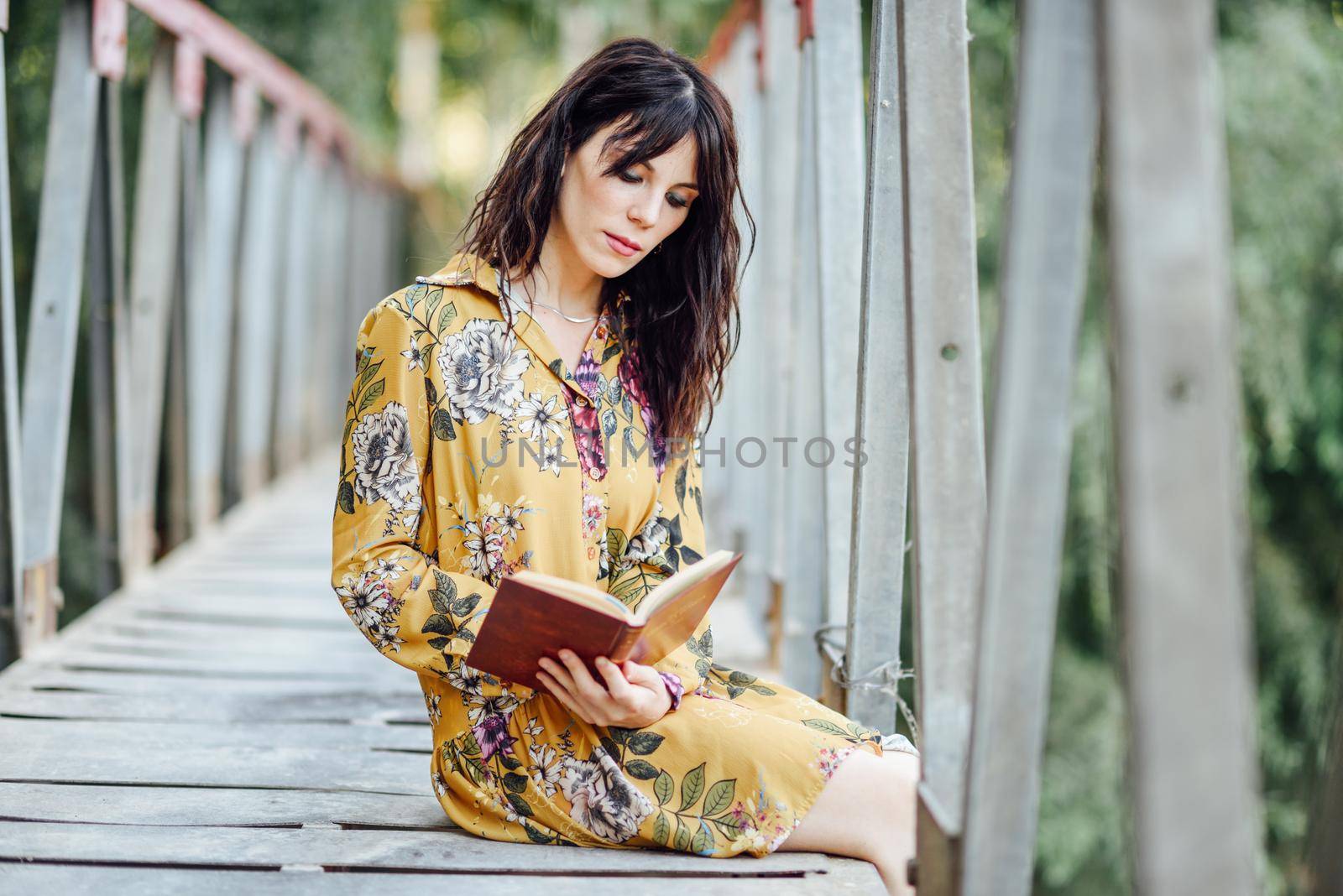 Young woman, wearing flowered dress, reading a book on a rural bridge.