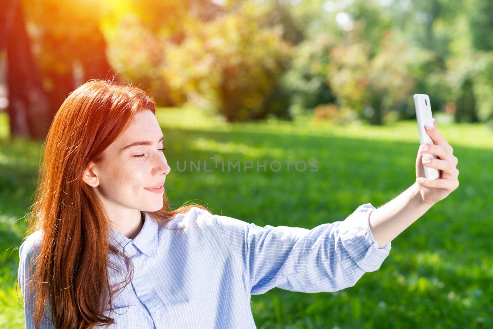 Young redhead woman taking selfie photo on green field. Close up portrait of charming girl with smartphone in hand. Lady having fun in spring park at sunny summer day. Digital mobile technology